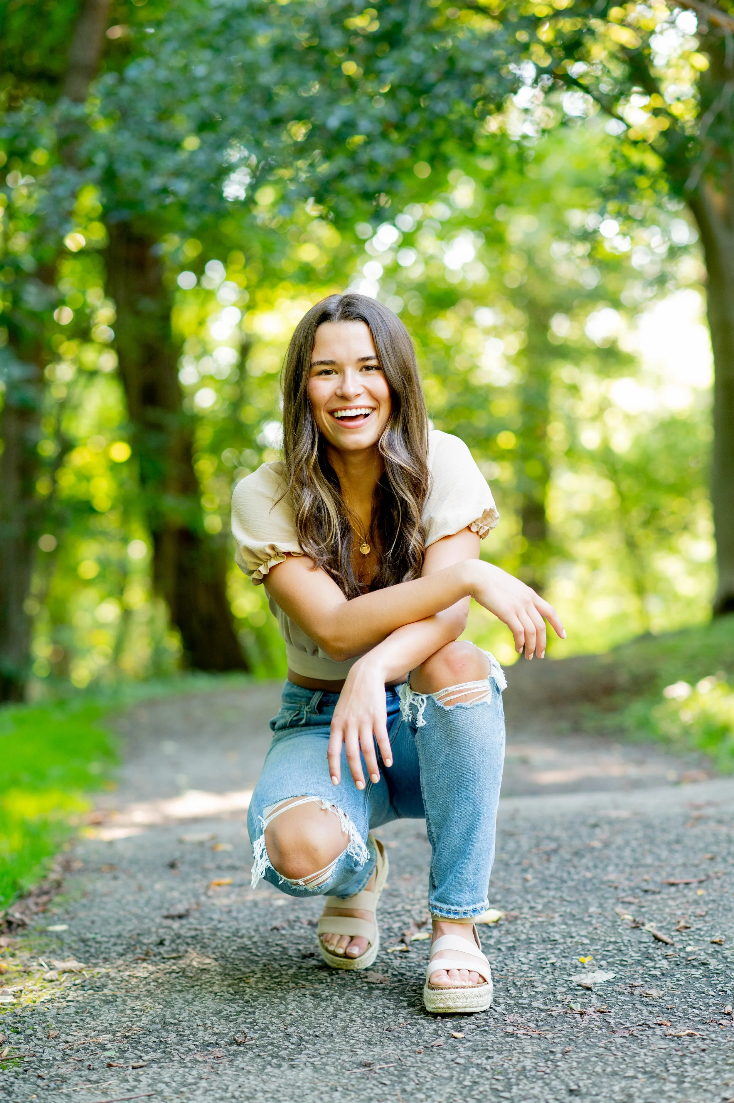 Pittsburgh Senior Photography.  A young woman with long brown hair, wearing a beige top and ripped jeans, crouching on a pathway in a lush green forest, smiling at the camera.
