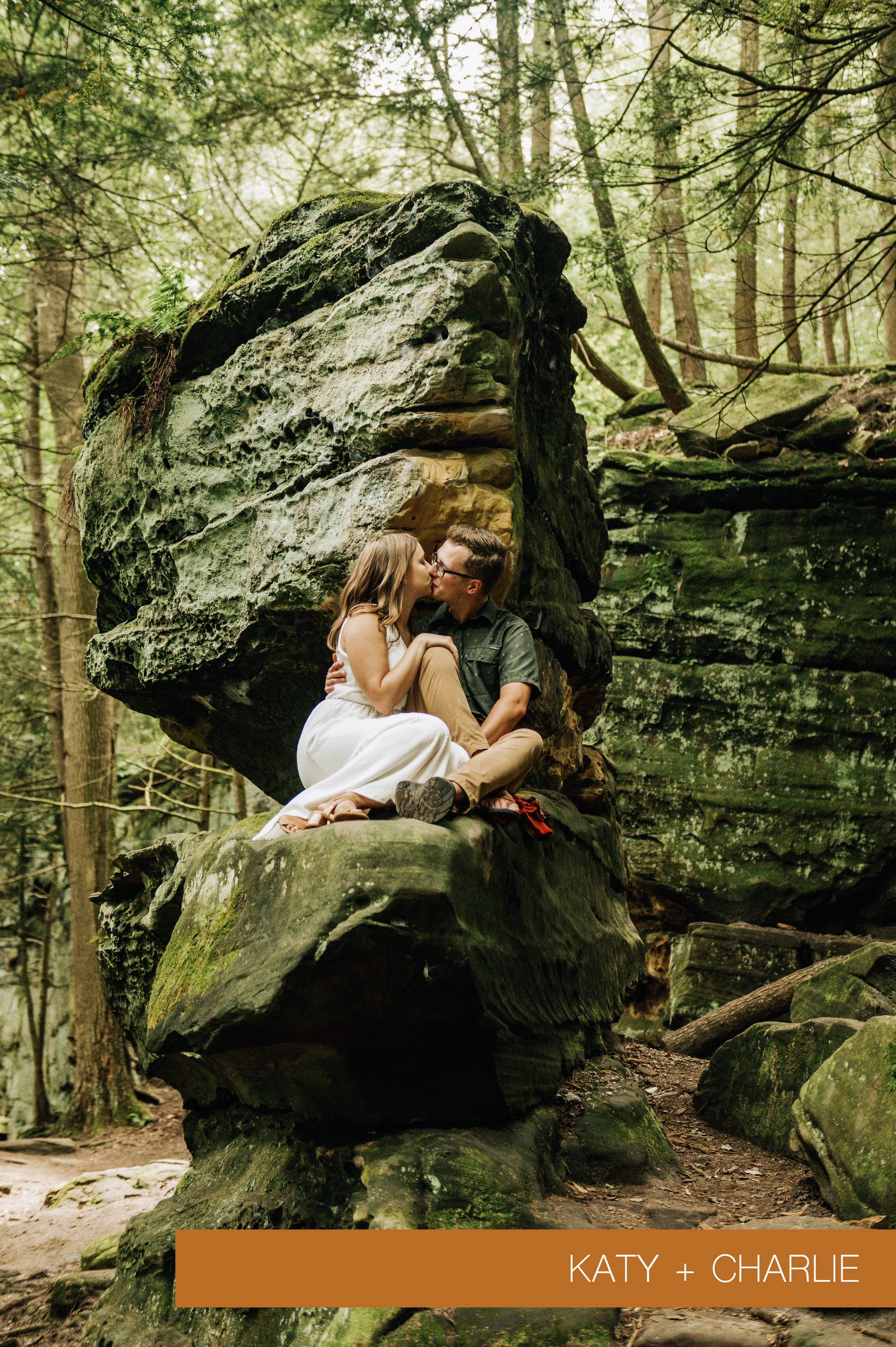A couple sitting on a rock in a forest, sharing a kiss. The woman is wearing a white dress and the man is in a green shirt and tan pants. Large moss-covered rock formations and trees surround them.
