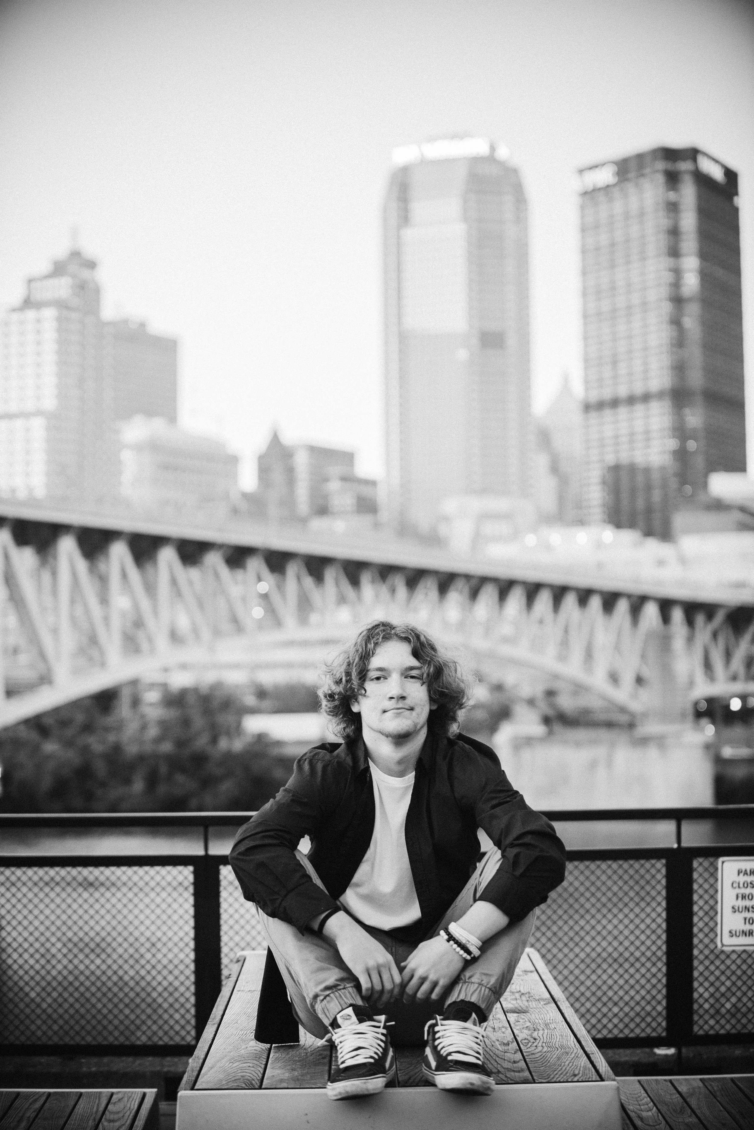 Pittsburgh Senior Photography.  A young man with curly hair sitting on a wooden platform on a rooftop with a city skyline and bridge in the background, black and white photo.