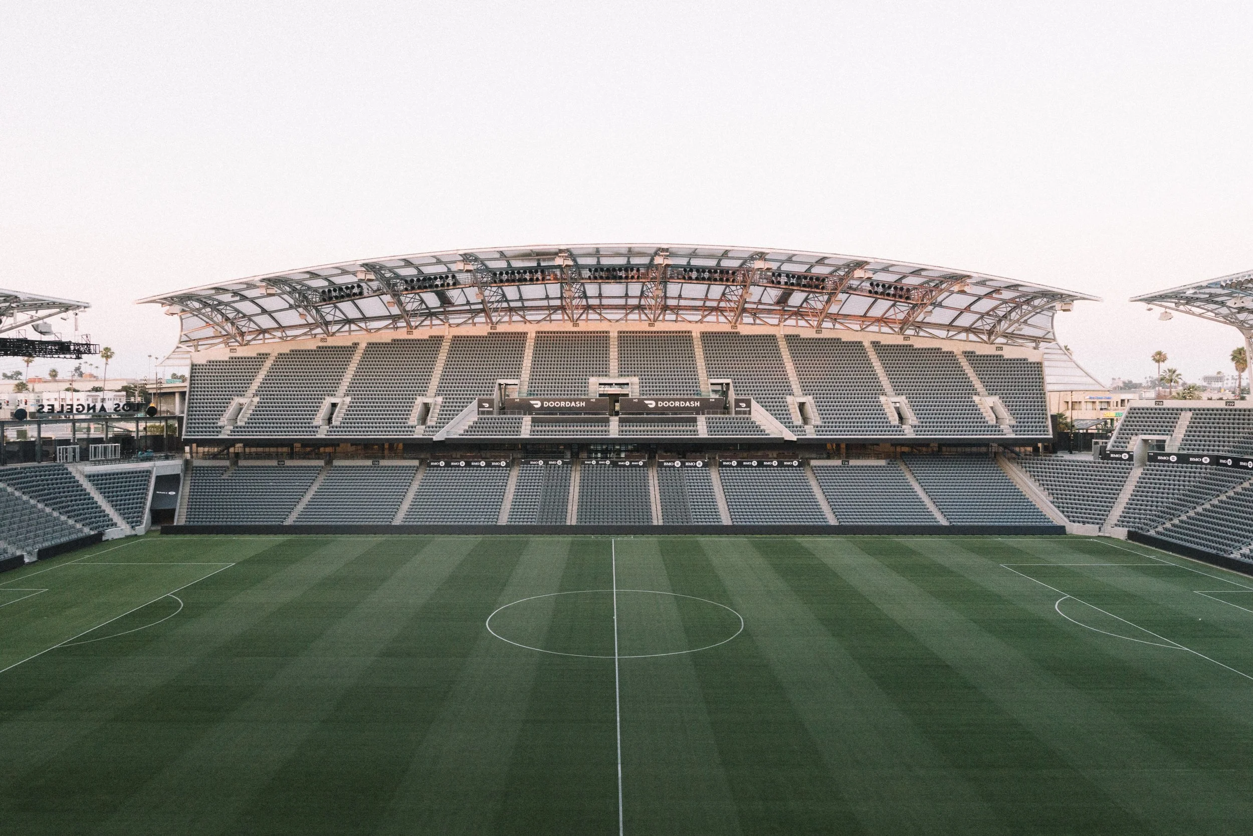 Empty soccer stadium with green field, seats, and a large covered section in the background, under a clear sky.