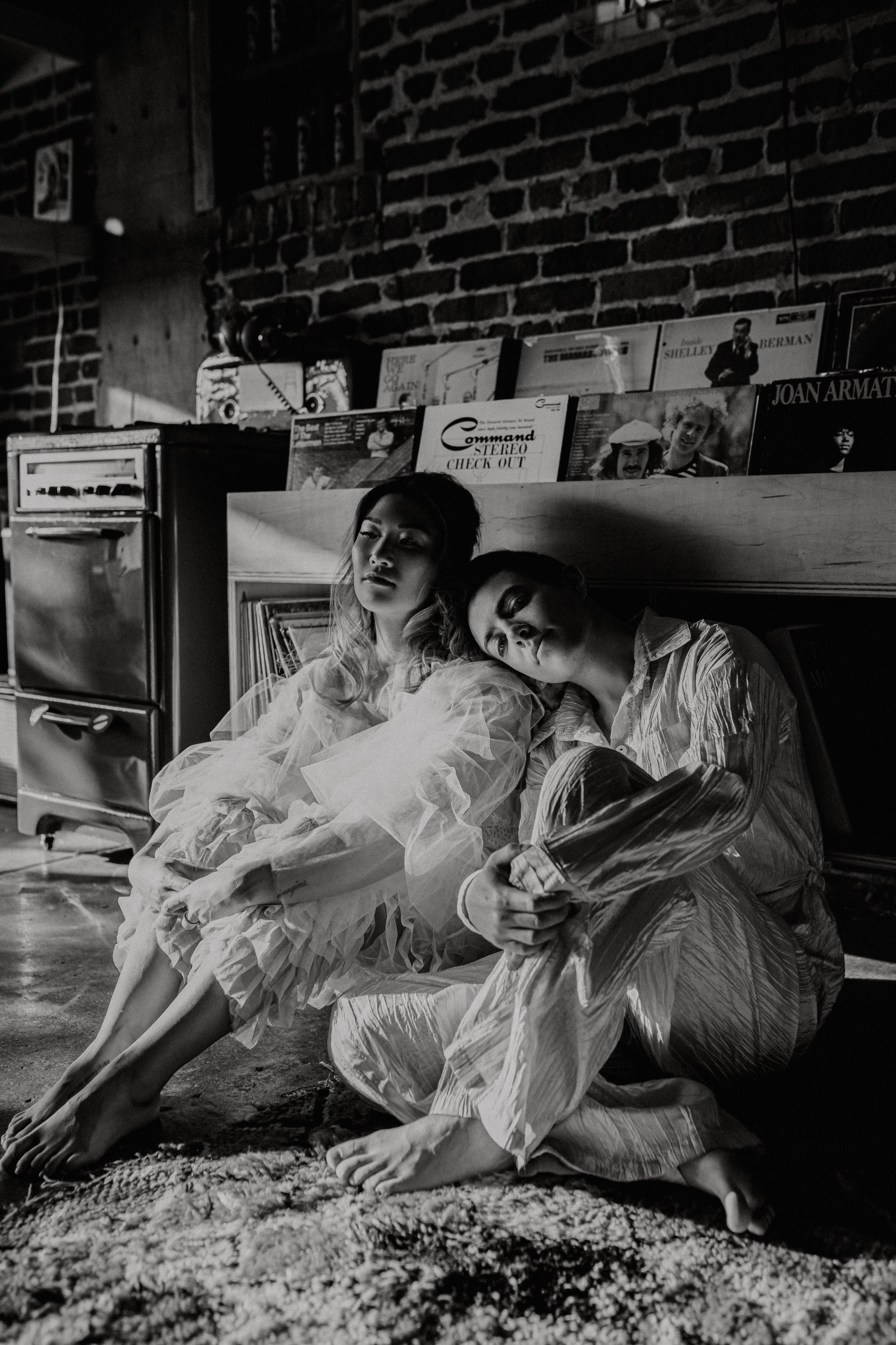 Black and white photo of two women sitting on the floor with their backs against a brick wall in a room filled with records. One woman has long hair and the other has shorter hair, both appear contemplative. There are shelves above them filled with v