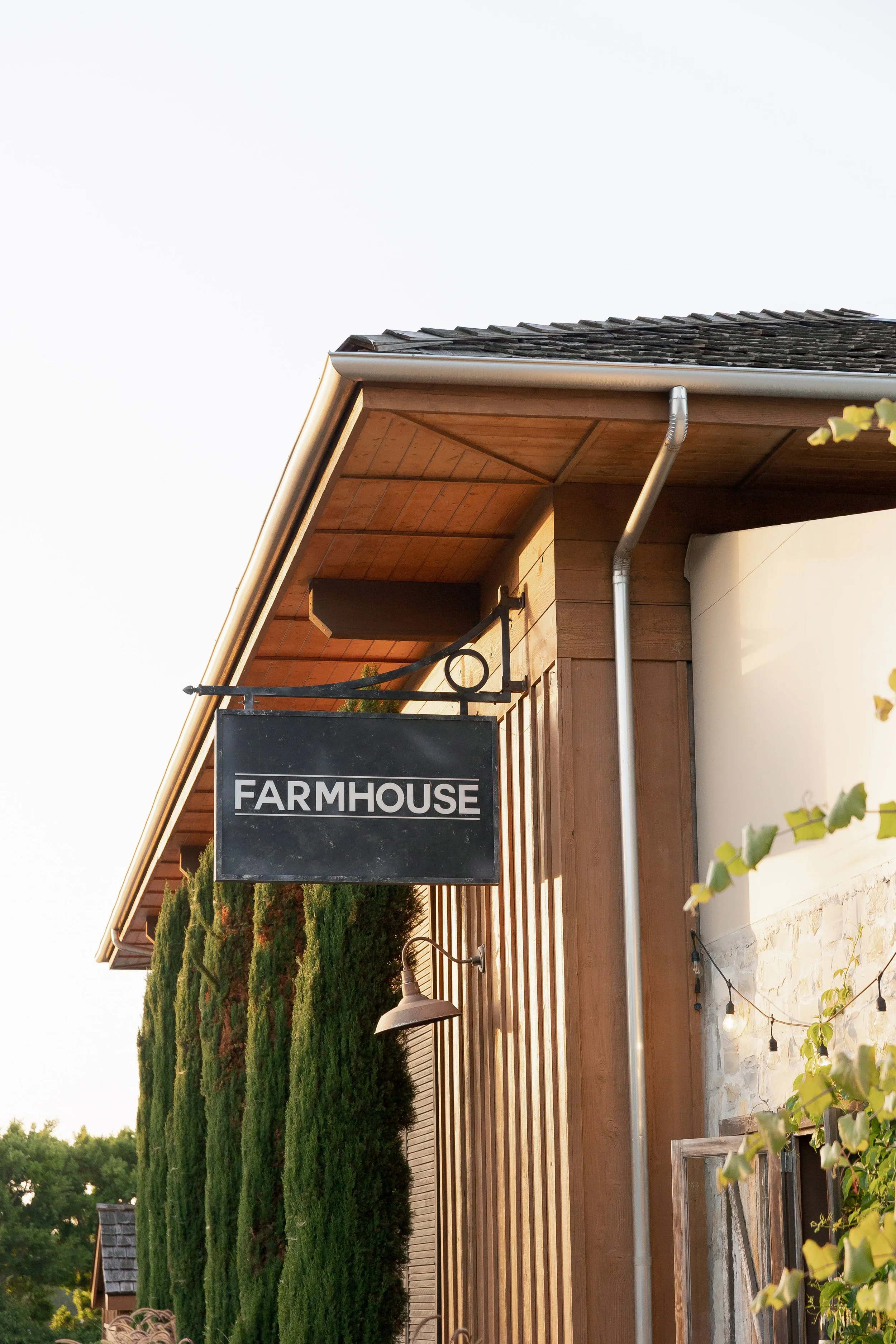 Exterior of a building with a modern design, featuring a sign that says 'Farmhouse,' a sloped roof with tiles, wooden paneling, and greenery in the background.