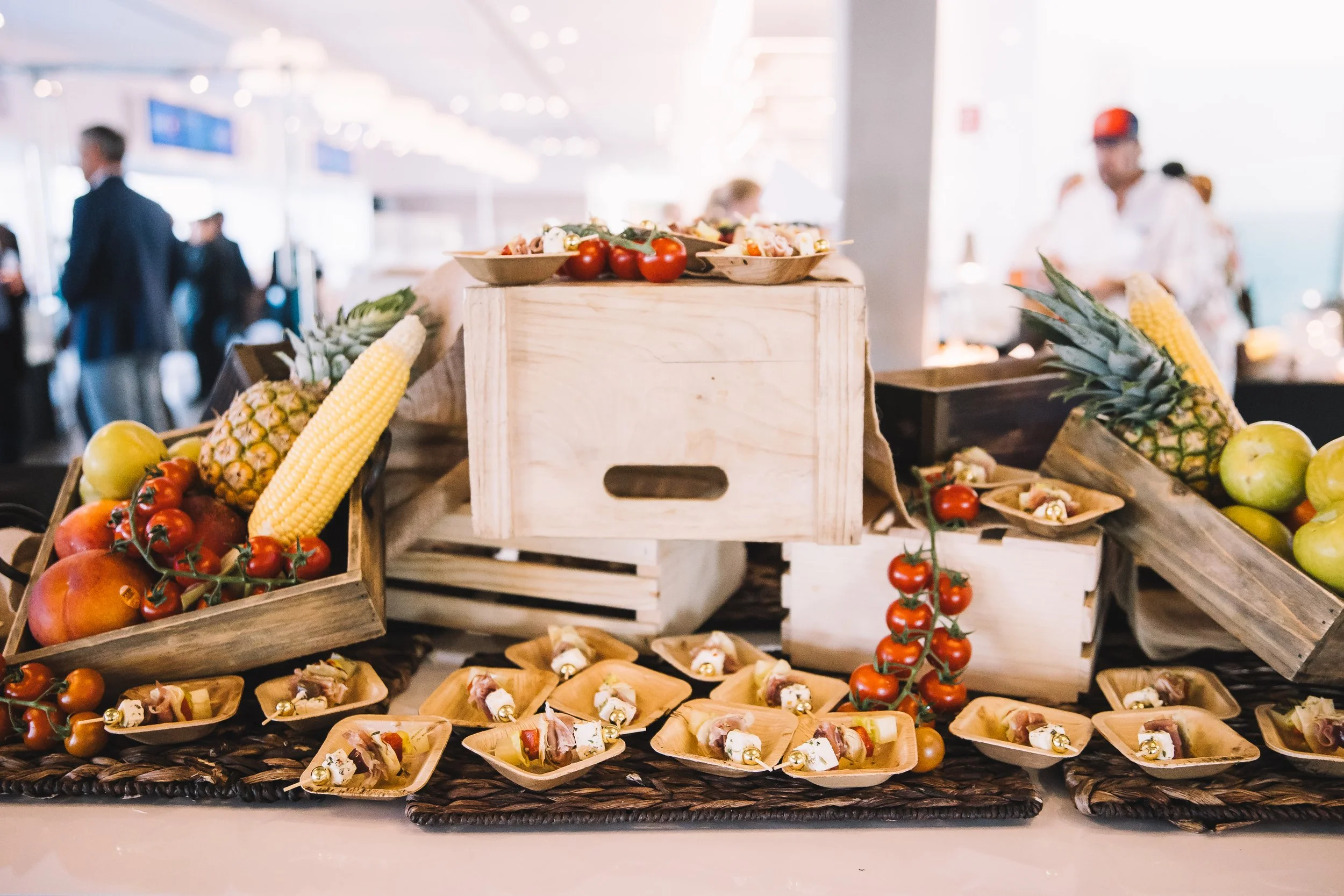 Display of fresh fruits such as pineapples, tomatoes, apples, and small appetizers arranged on wooden crates and on a table.