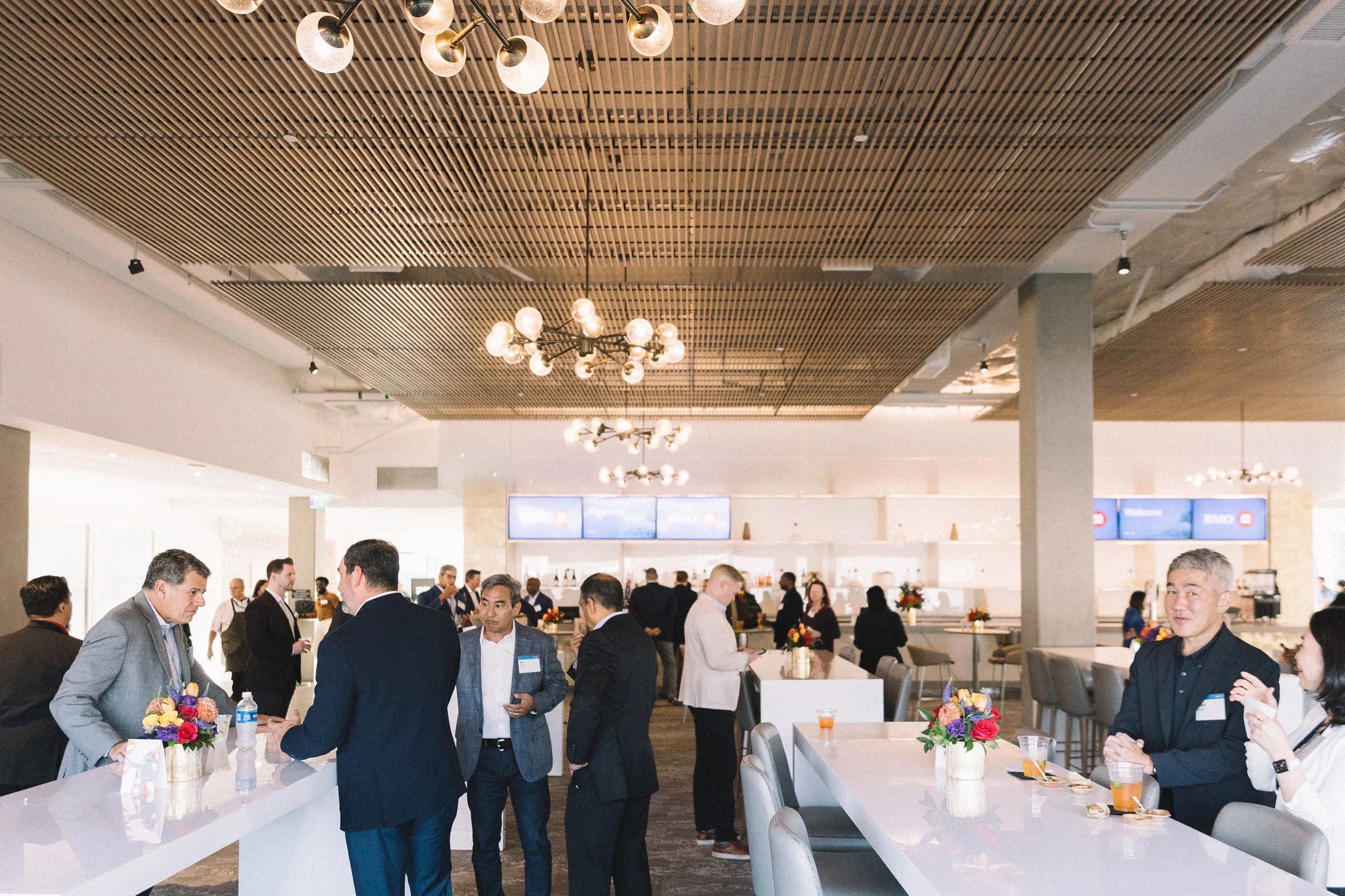 A busy indoor event with people in business attire, some holding drinks, and tables decorated with colorful flower arrangements. The ceiling features modern lighting fixtures, and there are electronic screens and a bar area in the background.