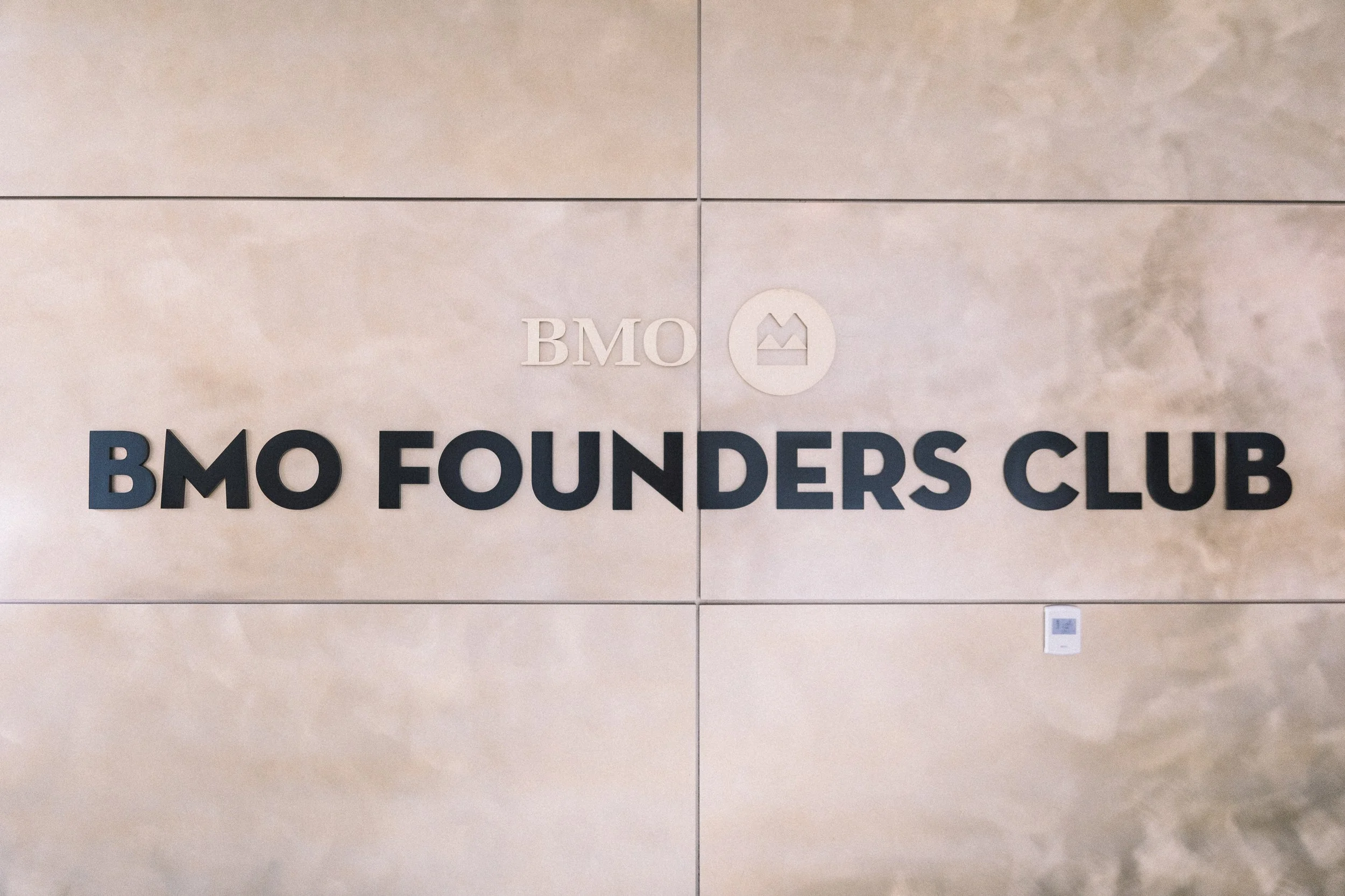 Close-up of a wall with the logo and text "BMO Founders Club" on it, featuring the BMO logo with a mountain and sun icon.
