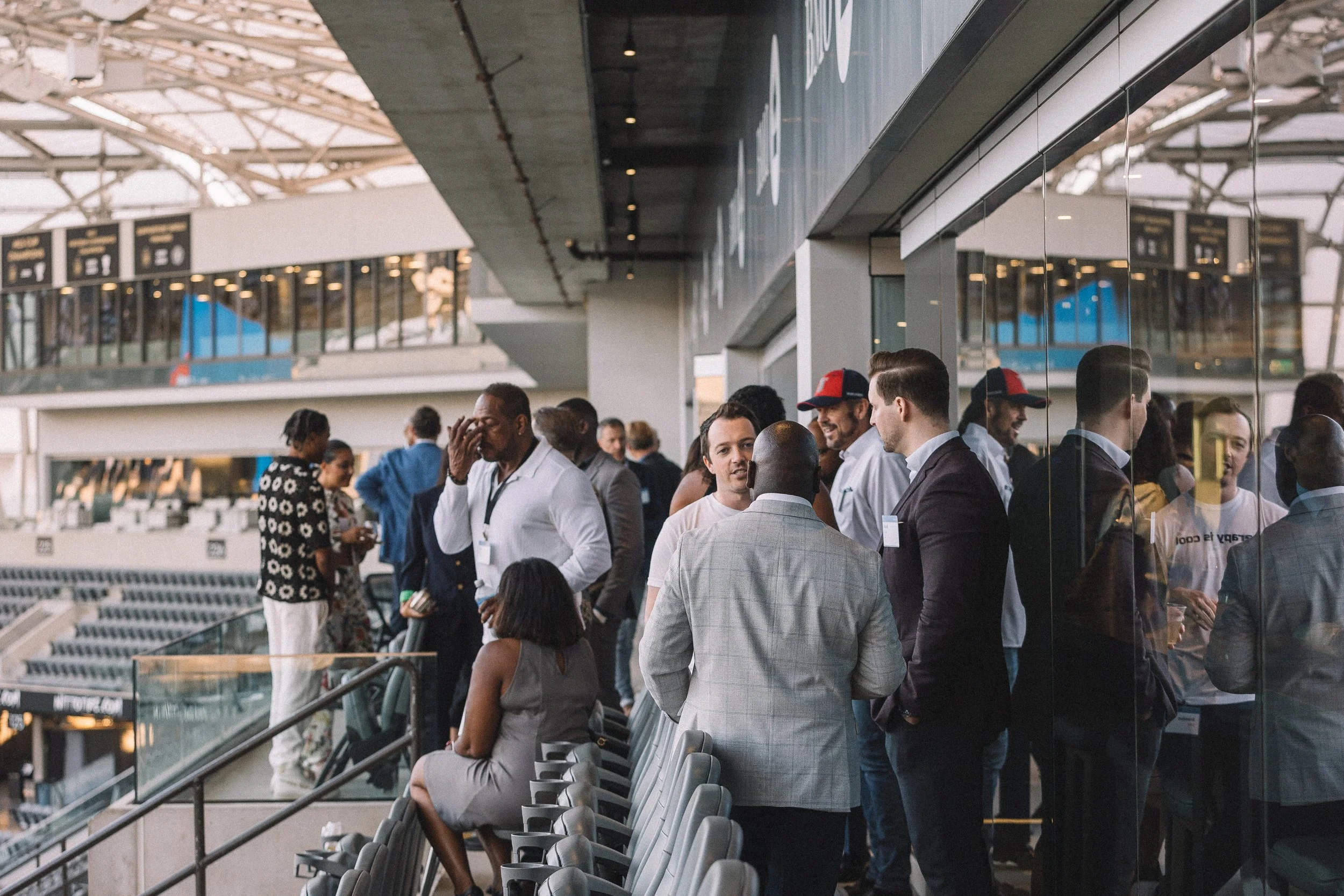 People waiting and socializing at an airport terminal, with some sitting and others standing near a reflective glass wall.