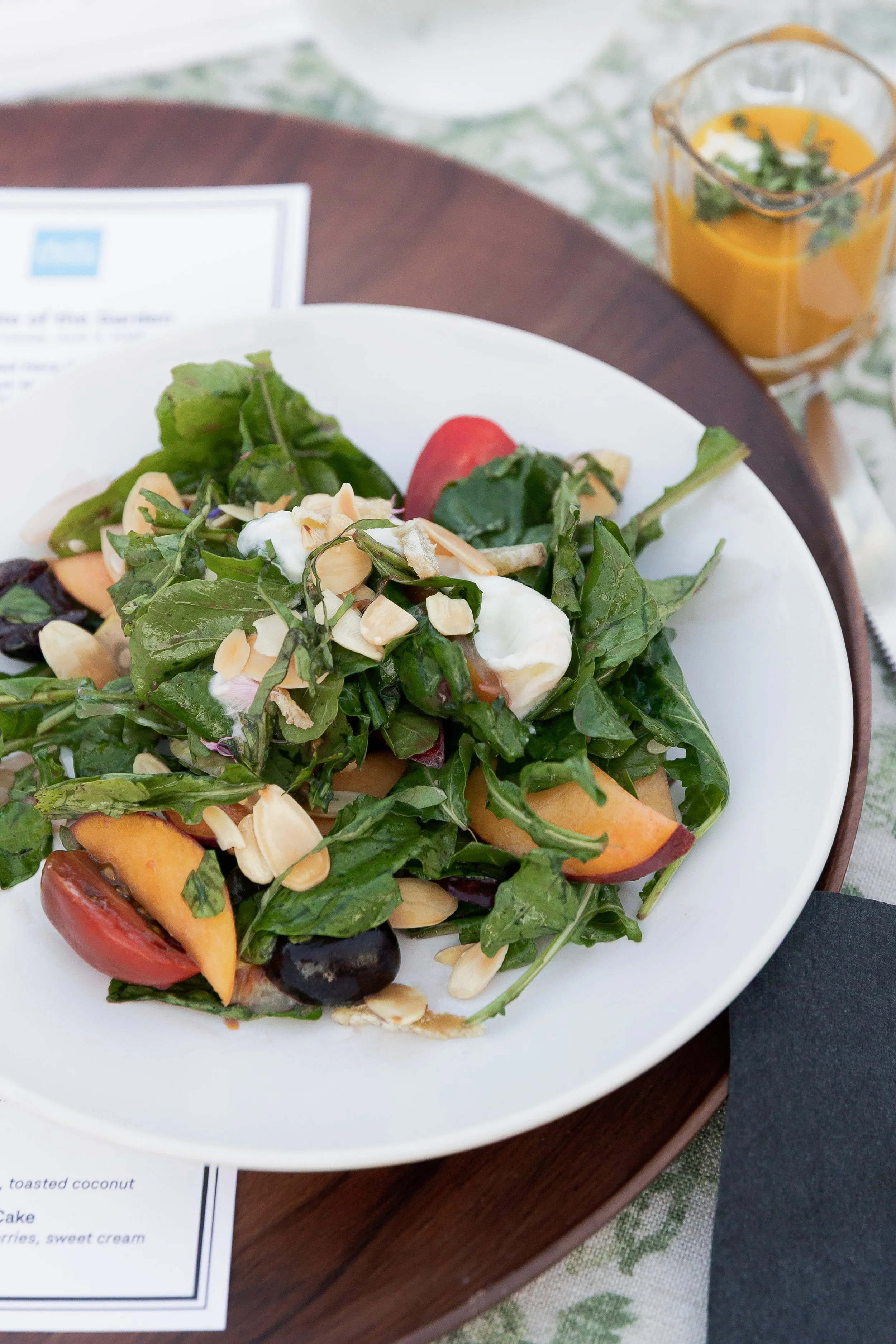A fresh green salad with sliced tomatoes, toasted coconut, and slivered almonds served on a white plate. A glass of orange juice with mint garnish is placed in the background.