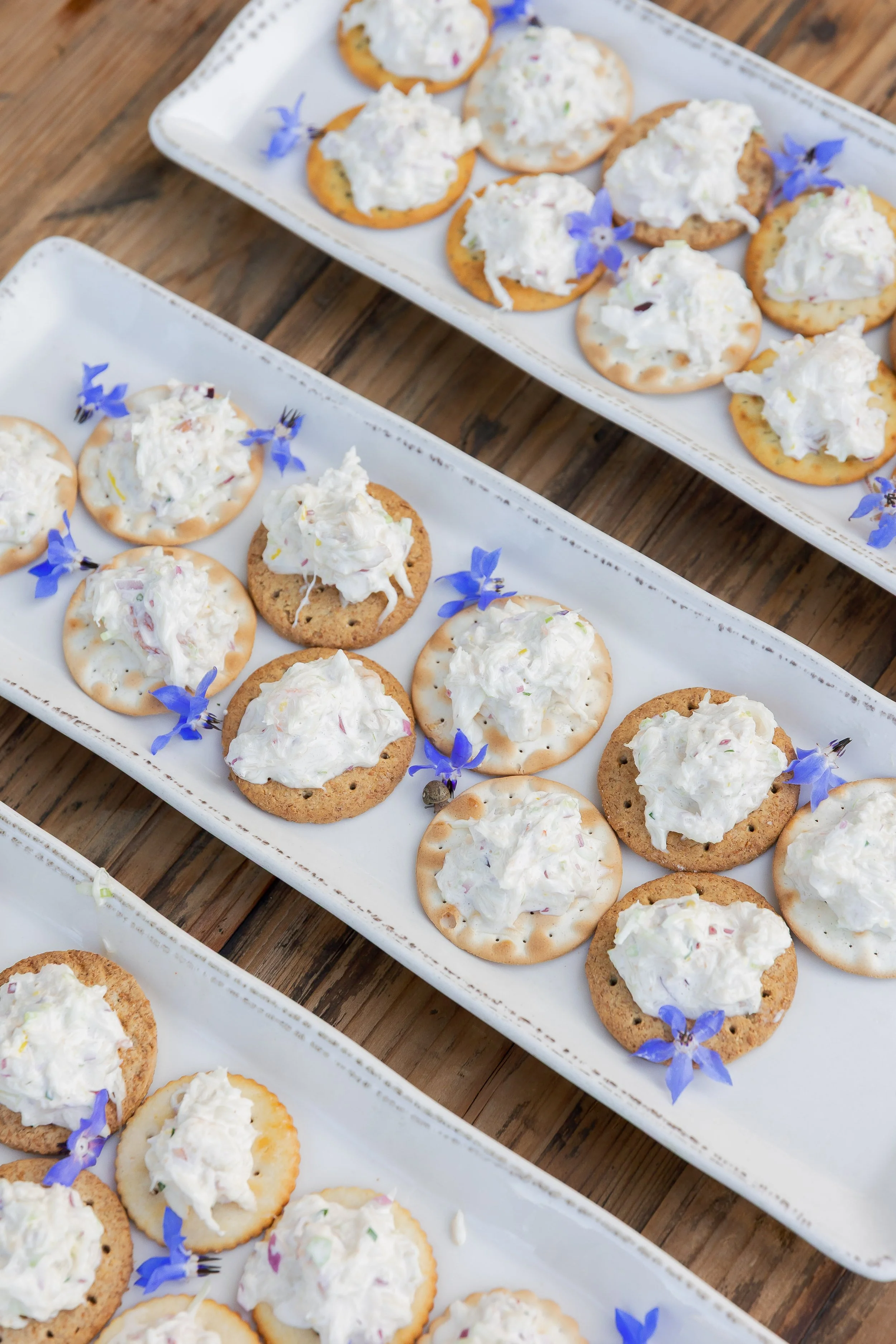 Plate of crackers topped with a white creamy spread, garnished with small purple flowers on a wooden surface.