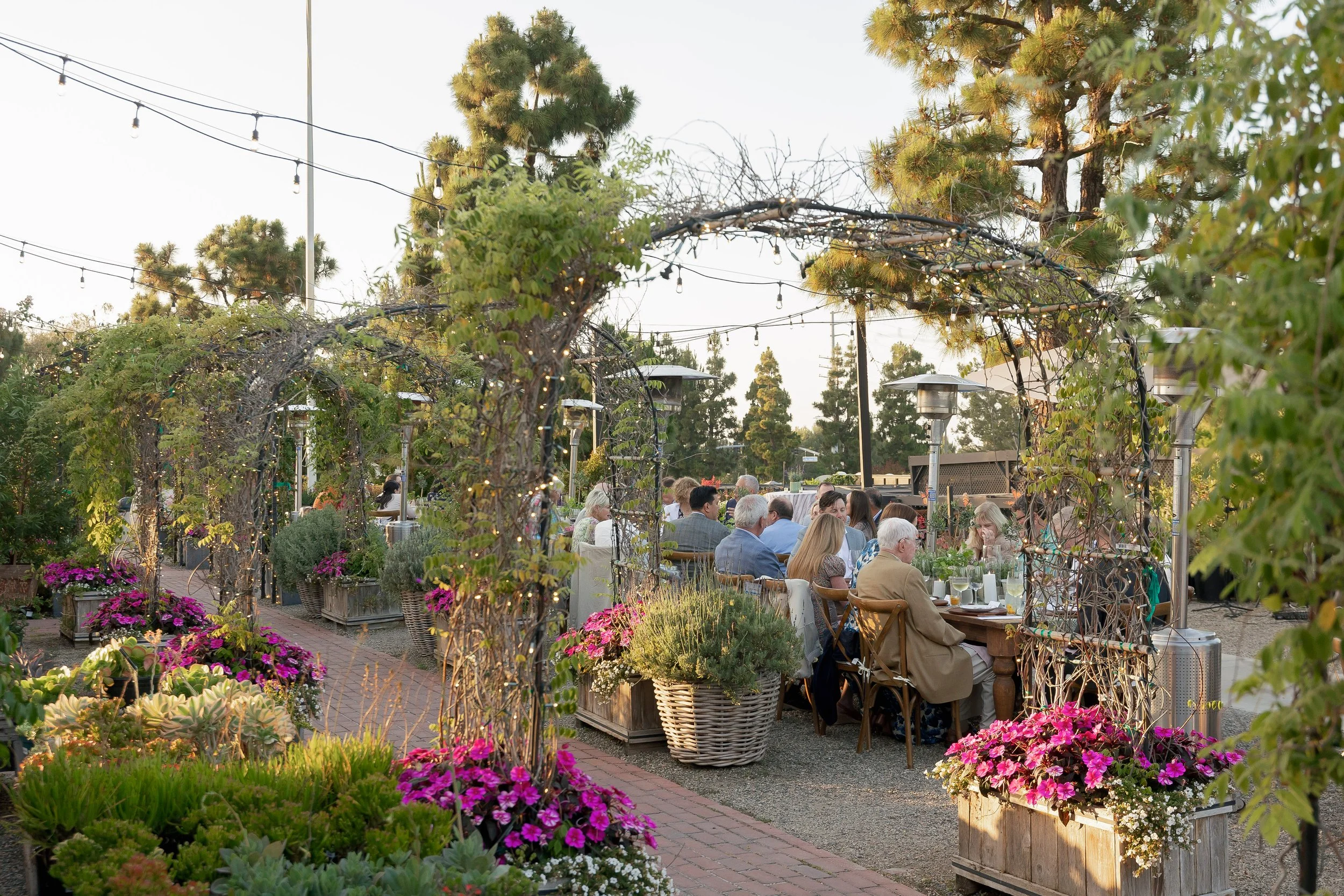 People dining at tables in an outdoor garden setting with flowering plants, string lights, and trees, during early evening or sunset.
