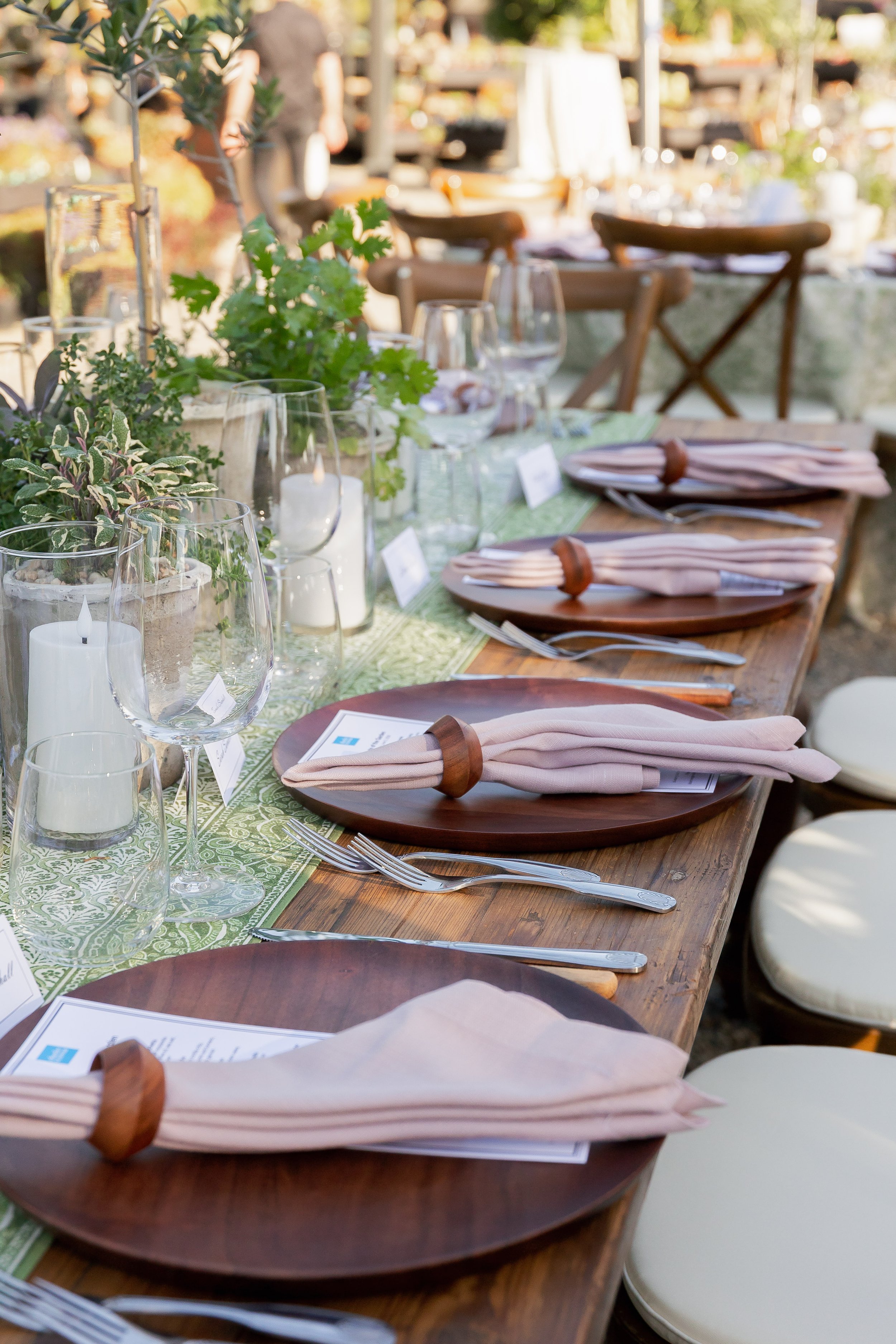 A long outdoor dinner table set with pink napkins, silverware, wine glasses, candles, and green plants, suggesting a prepared dining setup for an event or gathering.