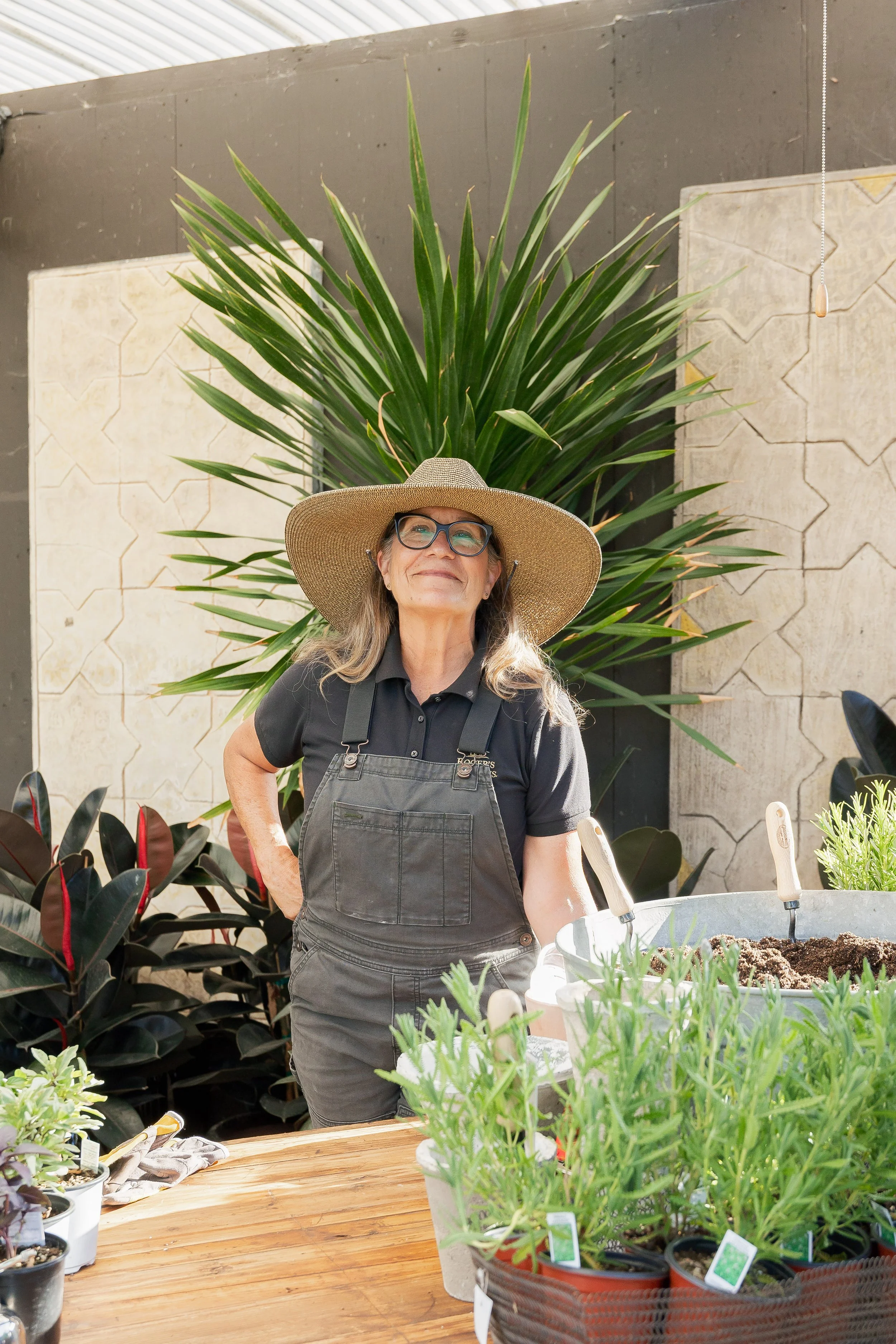 A woman wearing a wide-brimmed hat and glasses standing behind a table with potted plants in a gardening shop or nursery.