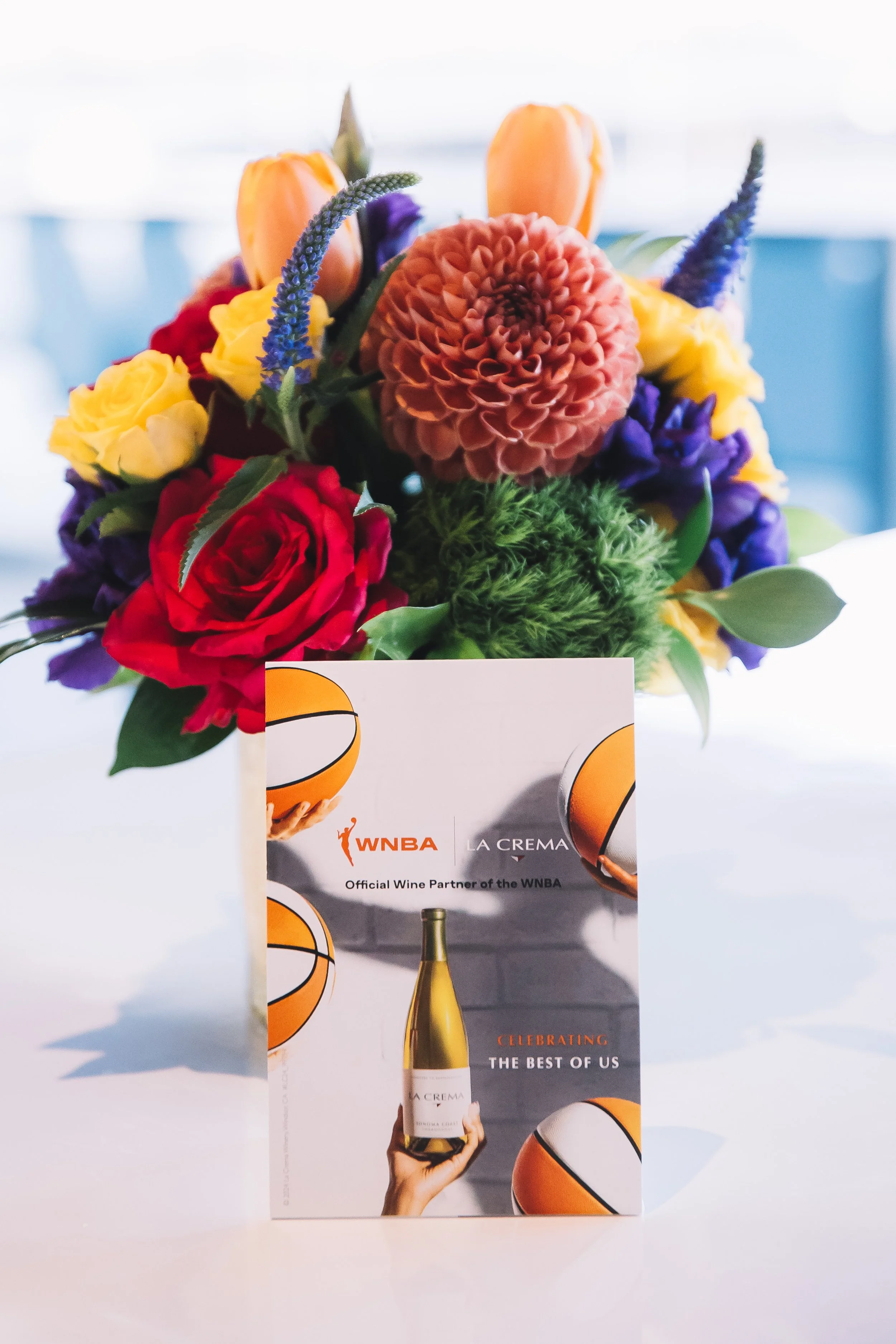 Vase with a colorful flower bouquet, including roses, dahlias, tulips, and delphiniums, placed next to a flyer celebrating the WNBA.