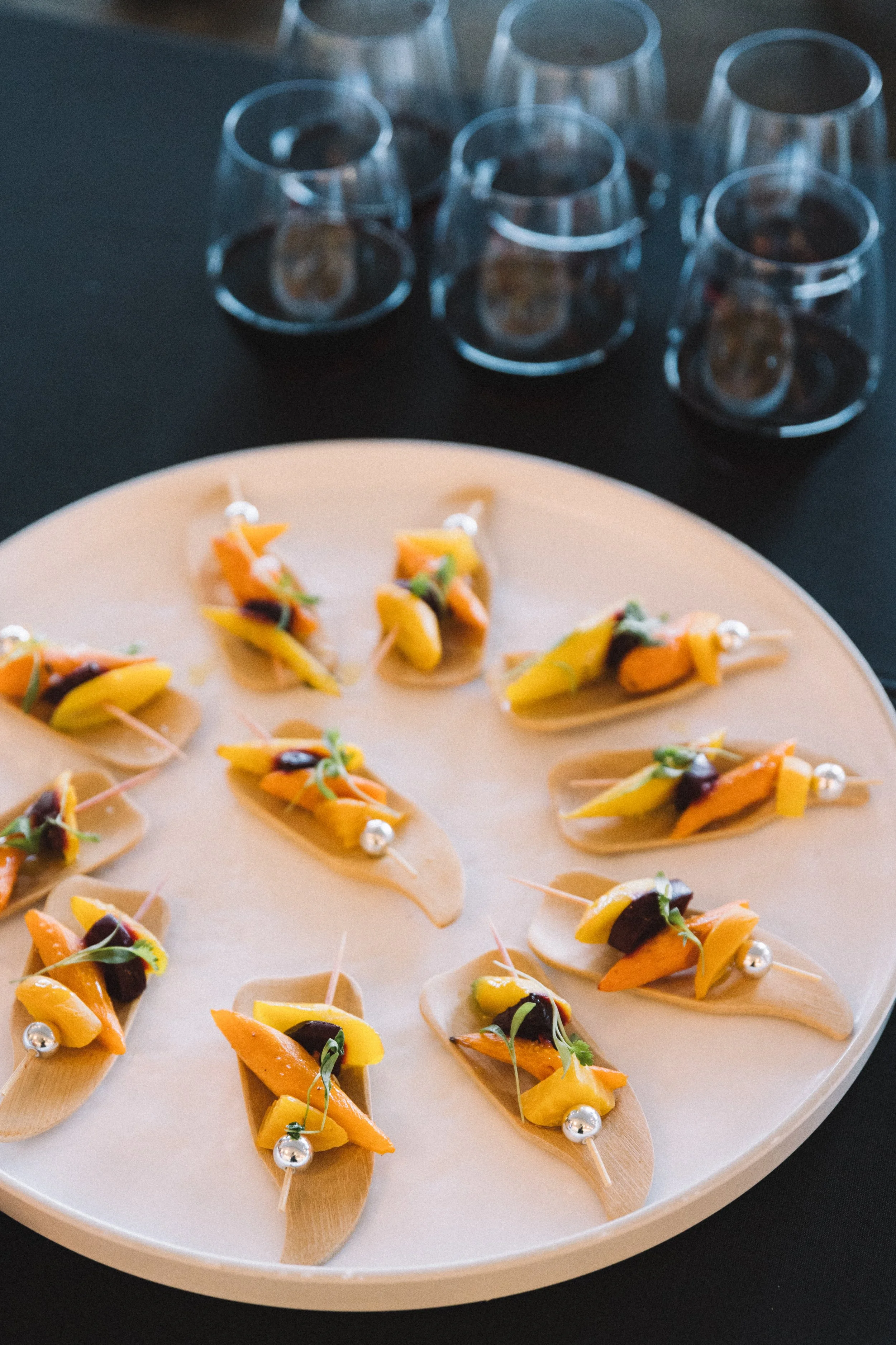 Appetizer bites on small wooden spoons with yellow and orange vegetables, garnished with microgreens and silver edible beads, arranged on a white circular platter.