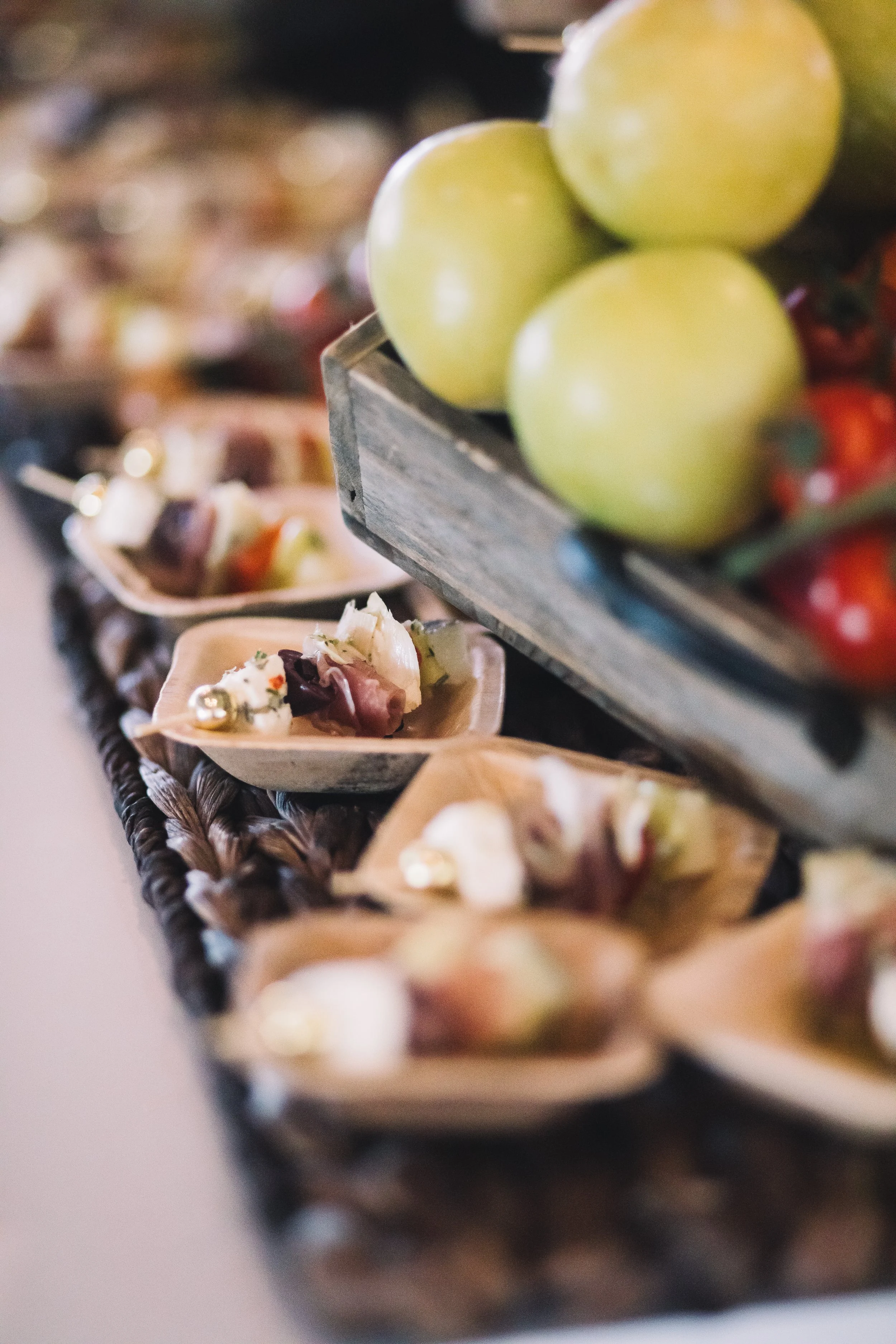Close-up of green grapes in a basket, with blurred background of assorted fruits and small wooden bowls with appetizers.