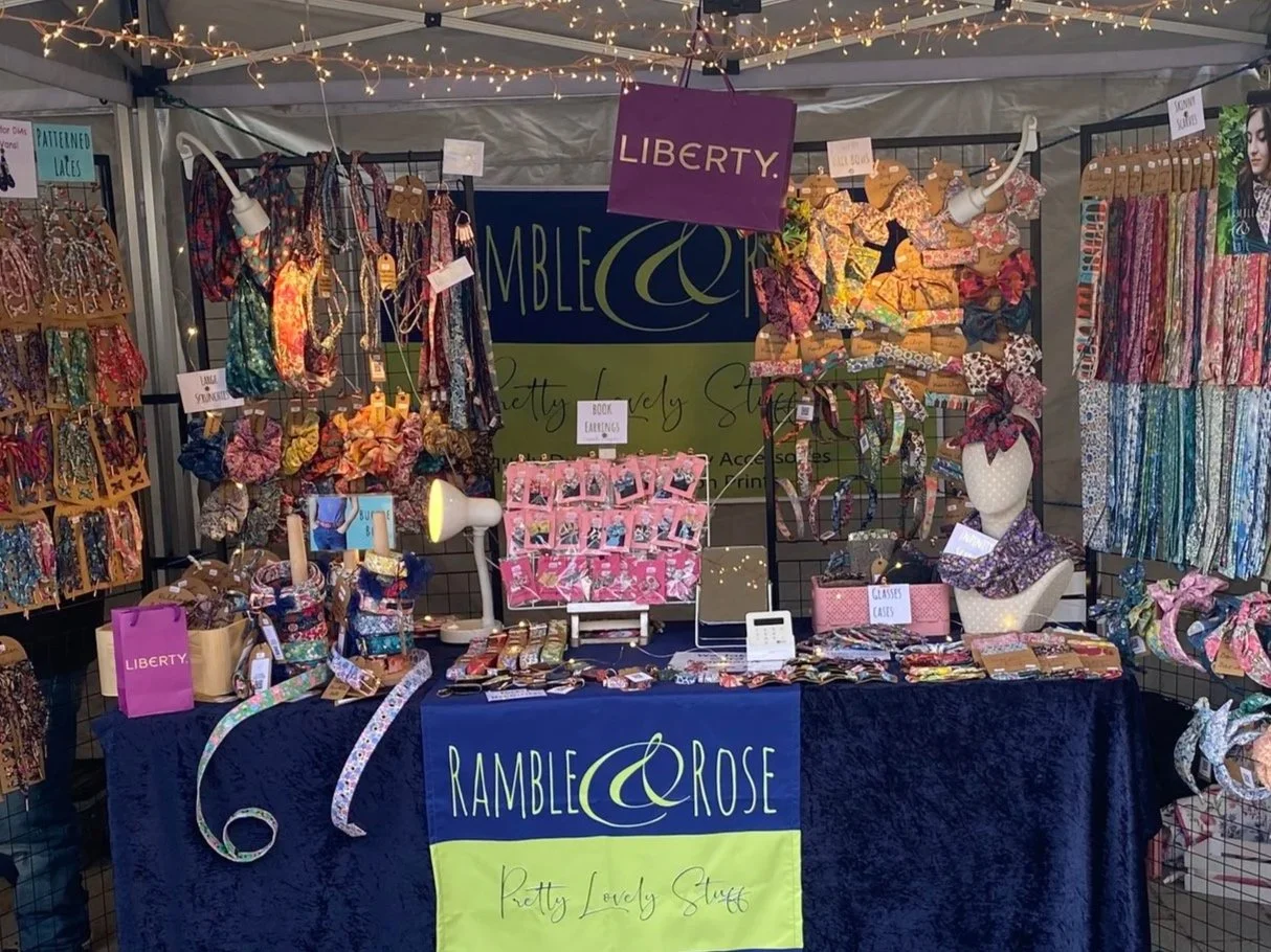 A booth at a craft fair displays colorful handmade jewelry, scarves, and fabric accessories. There are signs for 'Liberty,' 'Pretty Lovely Stuff,' and 'Book Earrings.' The booth has a blue tablecloth with a yellow and green banner, and string lights decorate the top.