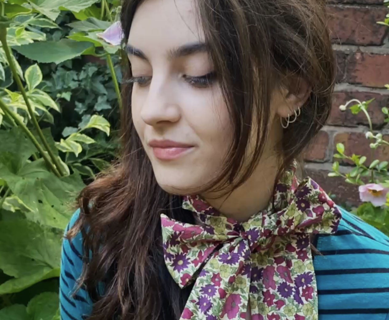 A young woman with long, wavy brown hair, wearing a colourful Liberty London Print  floral scarf tied around her neck, a blue and black striped shirt, and small hoop earrings, is standing outdoors amidst green plants and a brick wall background.