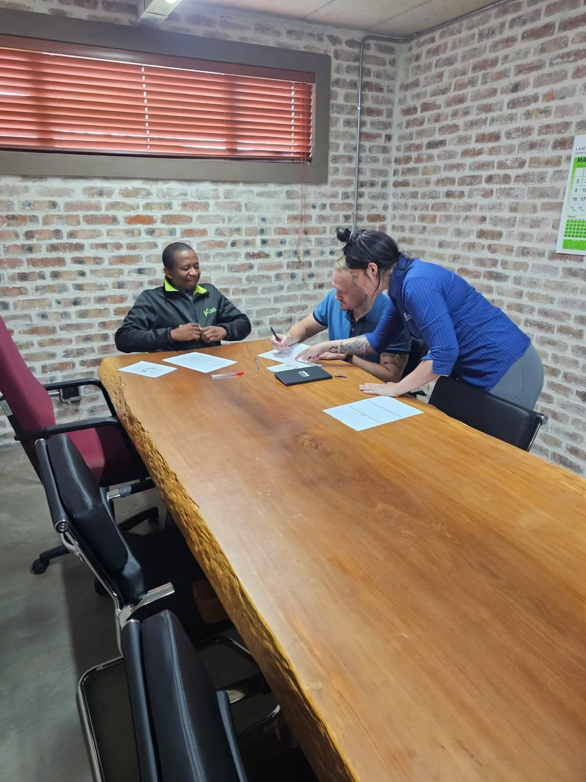 Three people sitting at a large wooden conference table in a room with brick walls. One person is sitting, and two are standing, engaged in a discussion or review of documents.