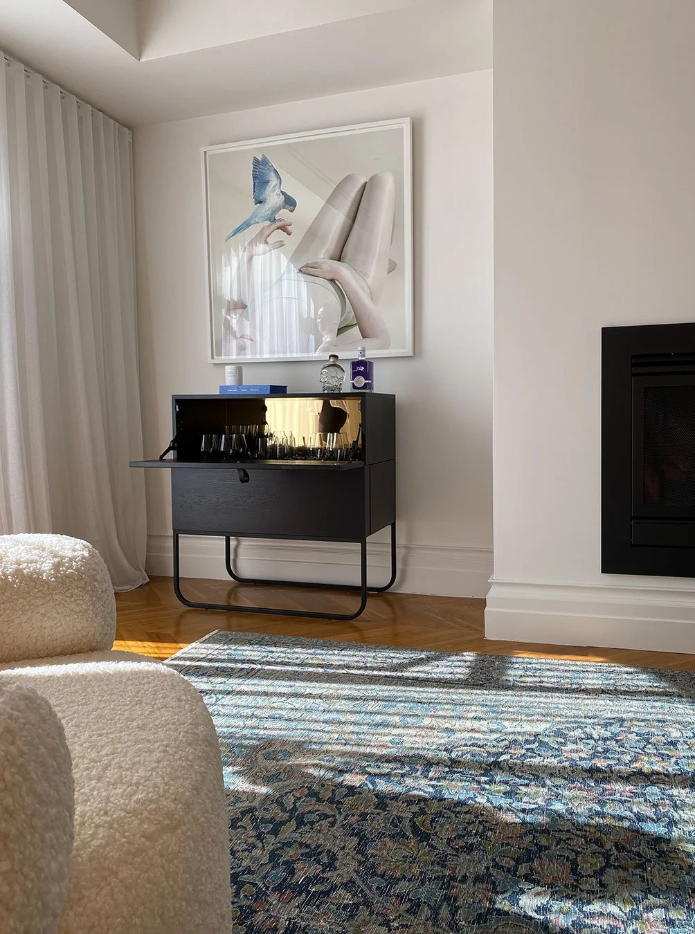 A living room with a black bar cart under a large framed artwork of a bird and a hand. The room has white walls, a hardwood floor, and a patterned area rug. Sunlight streams in, casting shadows.