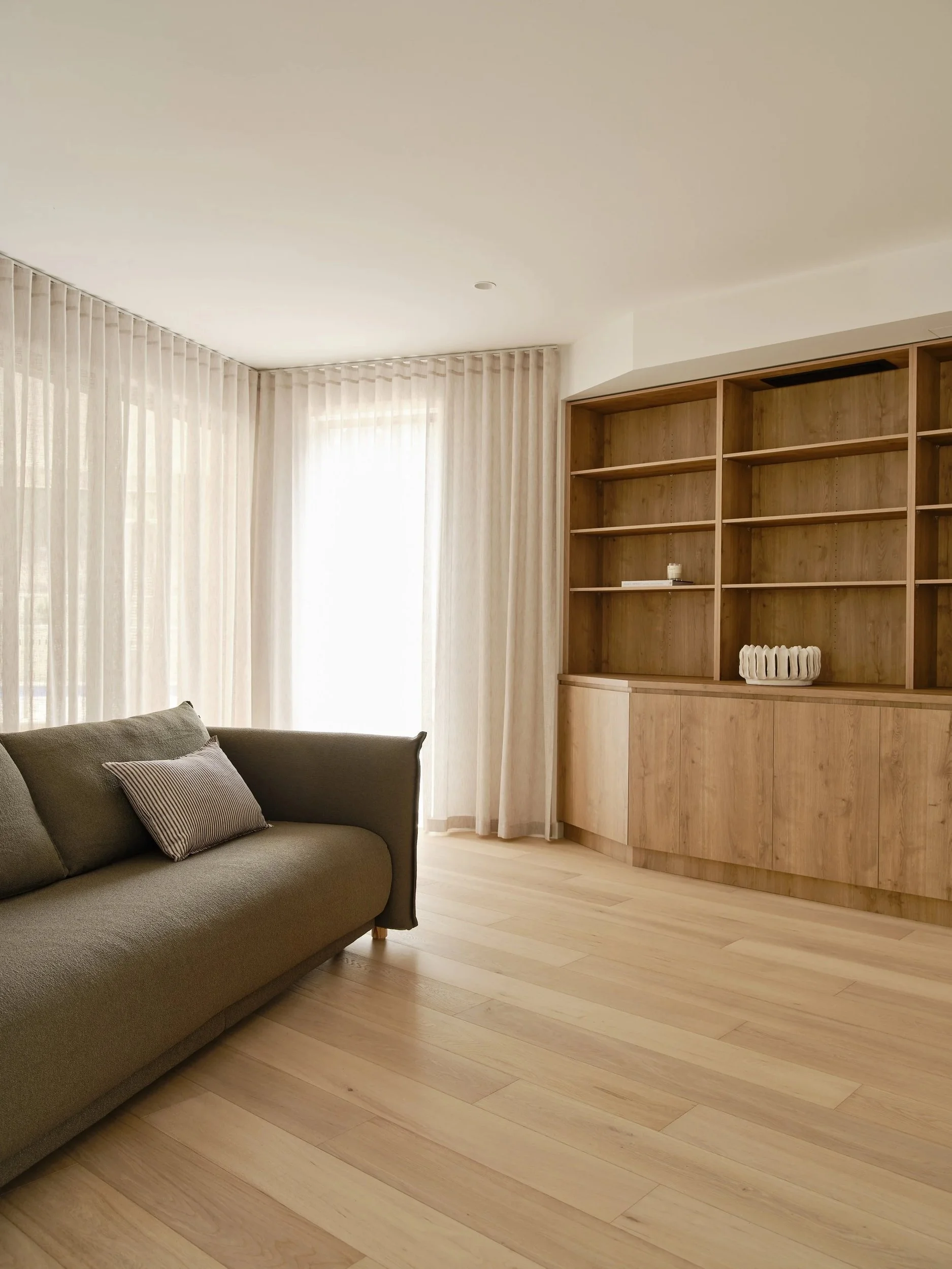 Sitting room with dark upholstered seating, full-height sheer curtains and timber display shelving.