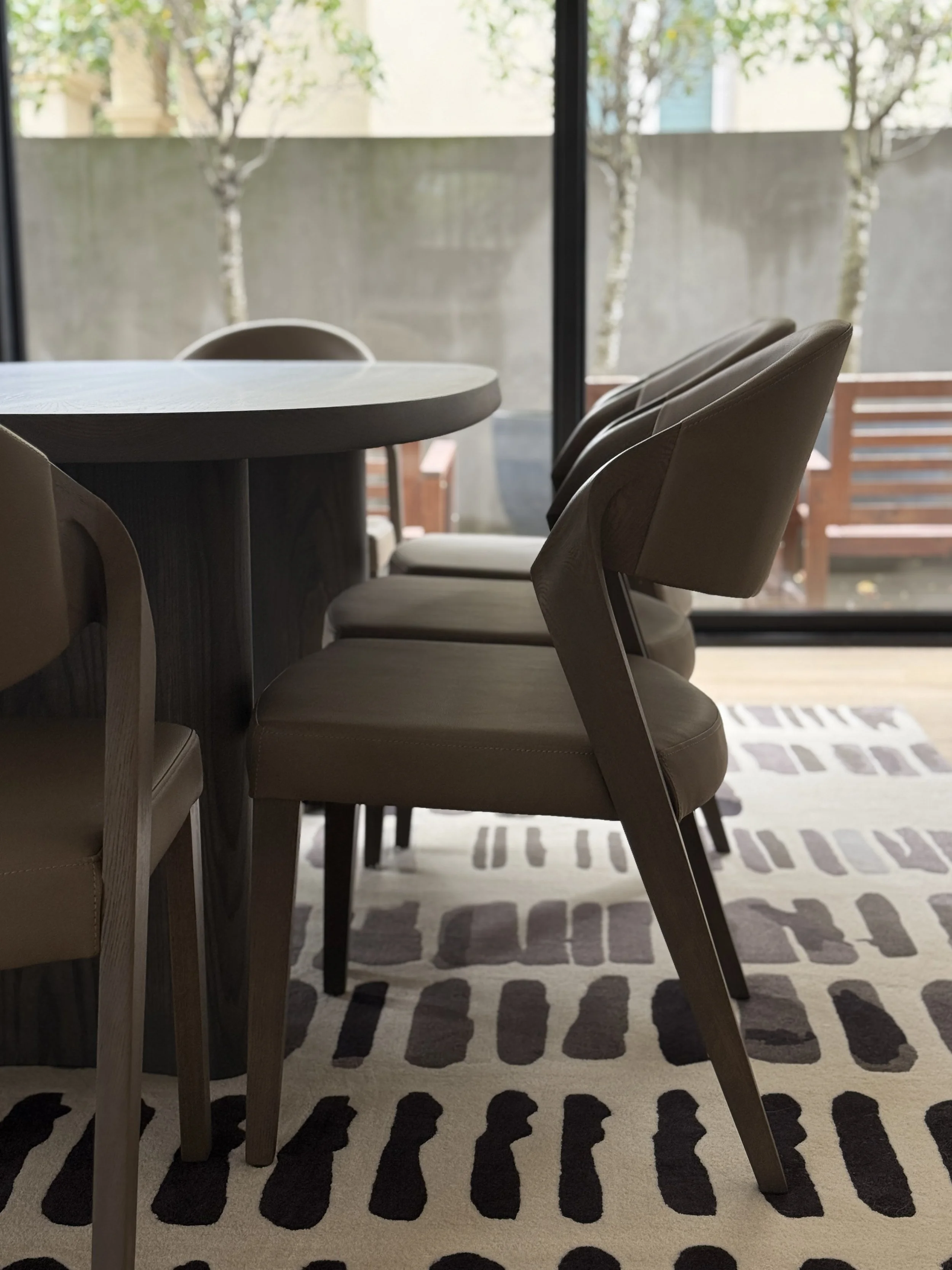 Interior of a dining area with a wooden table and beige chairs, a patterned rug on the floor, and large windows with a view of trees outside.