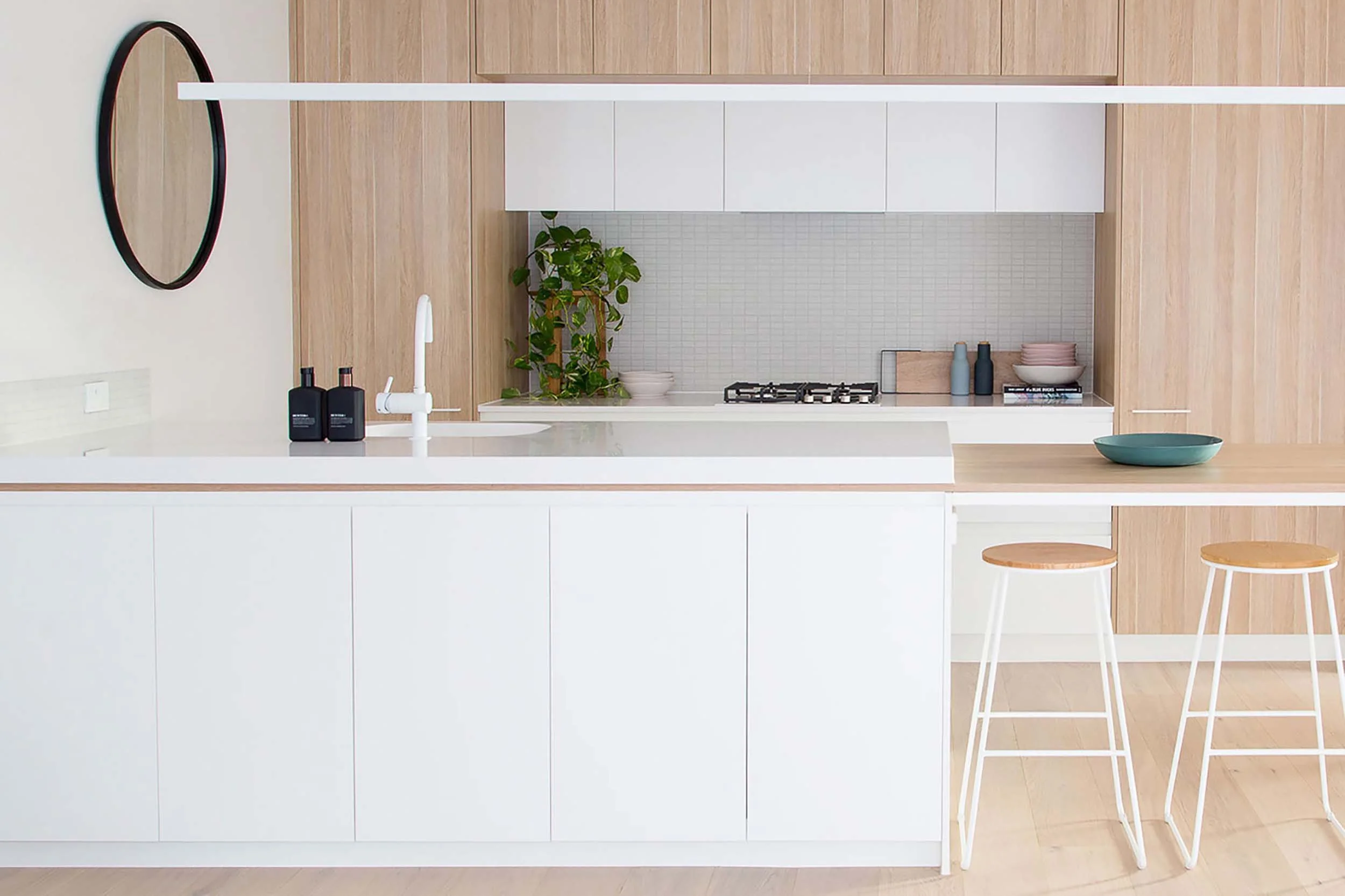 Modern kitchen with white countertops, wooden cabinets, a potted plant, and seating with two white chairs with wooden seats.