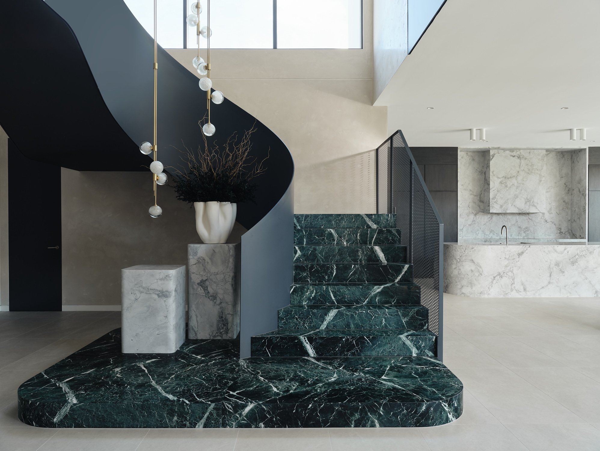 Green stone staircase with a curved dark balustrade and suspended globe lights opening onto the kitchen.