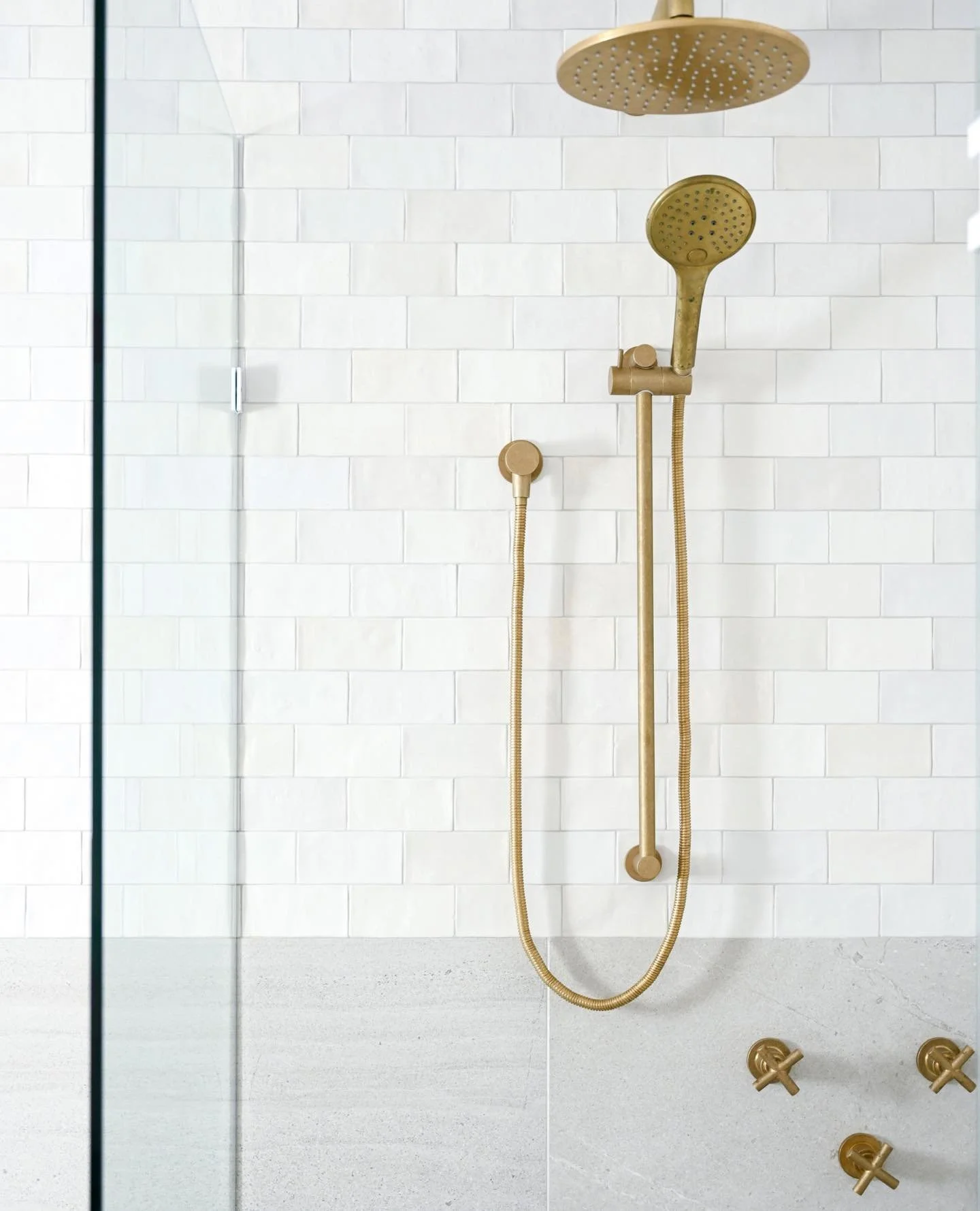 Modern shower with two gold shower heads, one large overhead and one handheld, against a white brick wall.