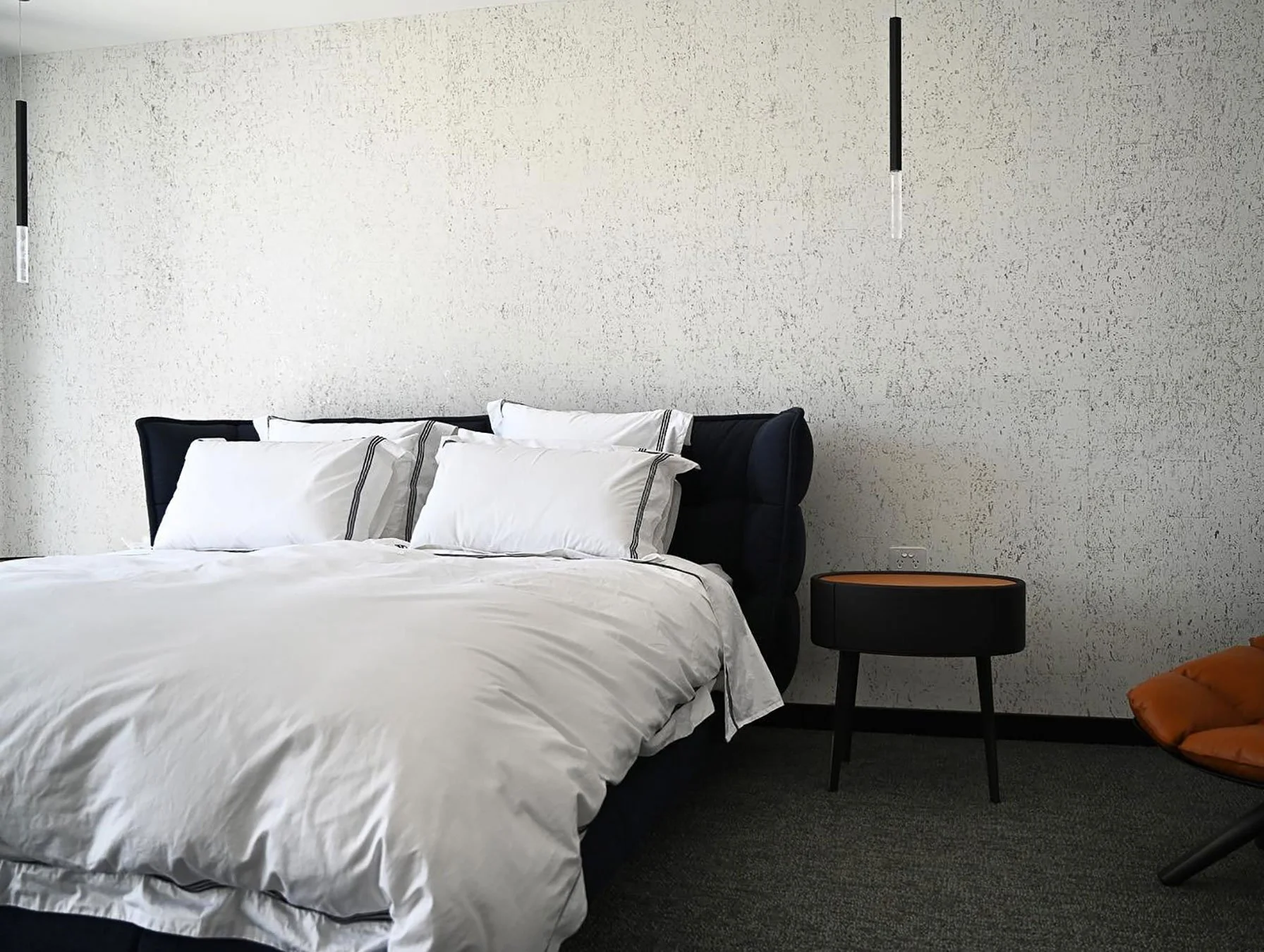 Modern bedroom with a bed featuring white linens and pillows, black upholstered headboard, and a side table next to the bed. The textured white wall and minimalist pendant lights add to the contemporary decor. An orange chair with a matching cushion 