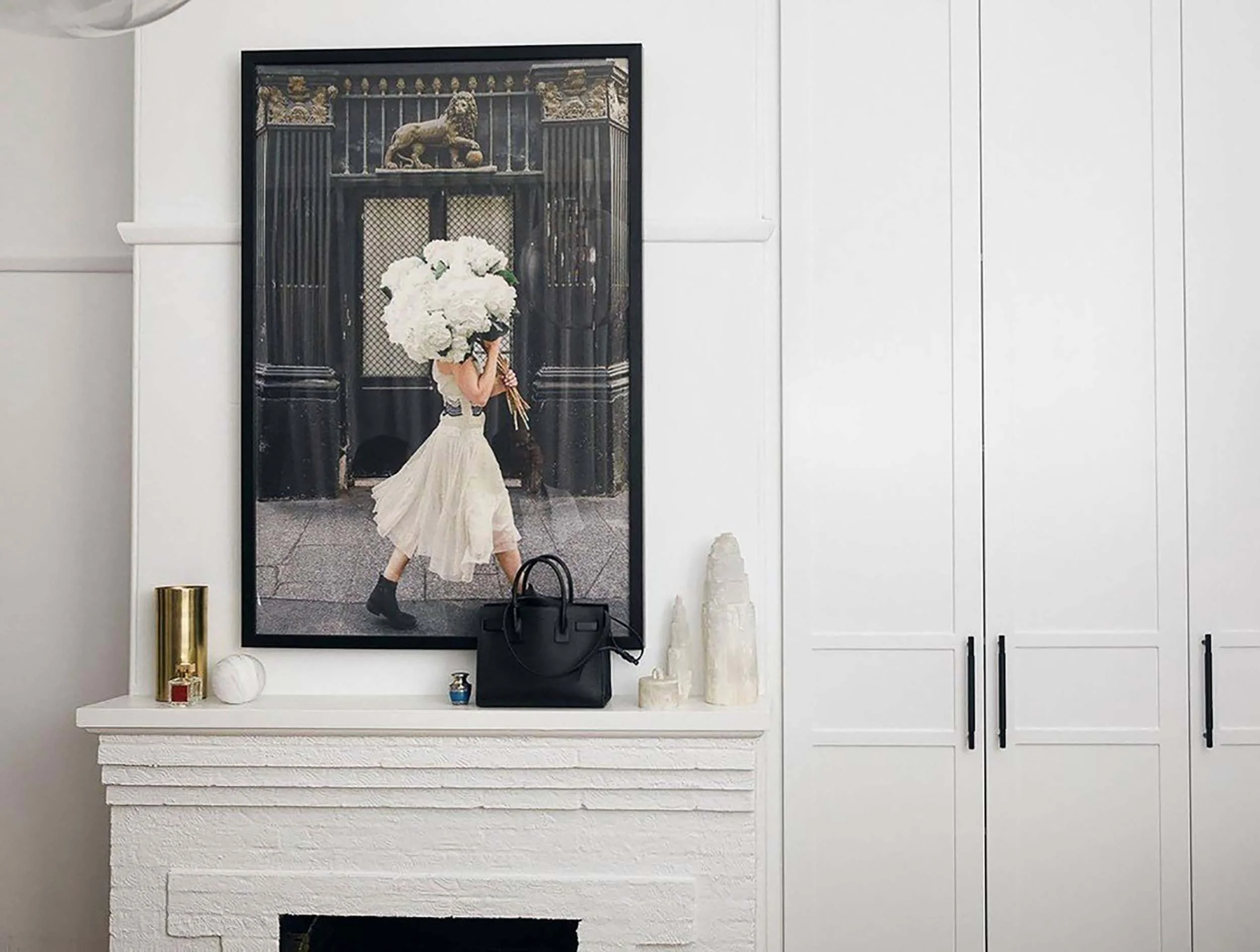 A reflection of a woman in a large decorative mirror with a black frame, holding a bouquet of white flowers. The scene includes a white stone fireplace mantel with decorative objects and a white wall with built-in cabinets.
