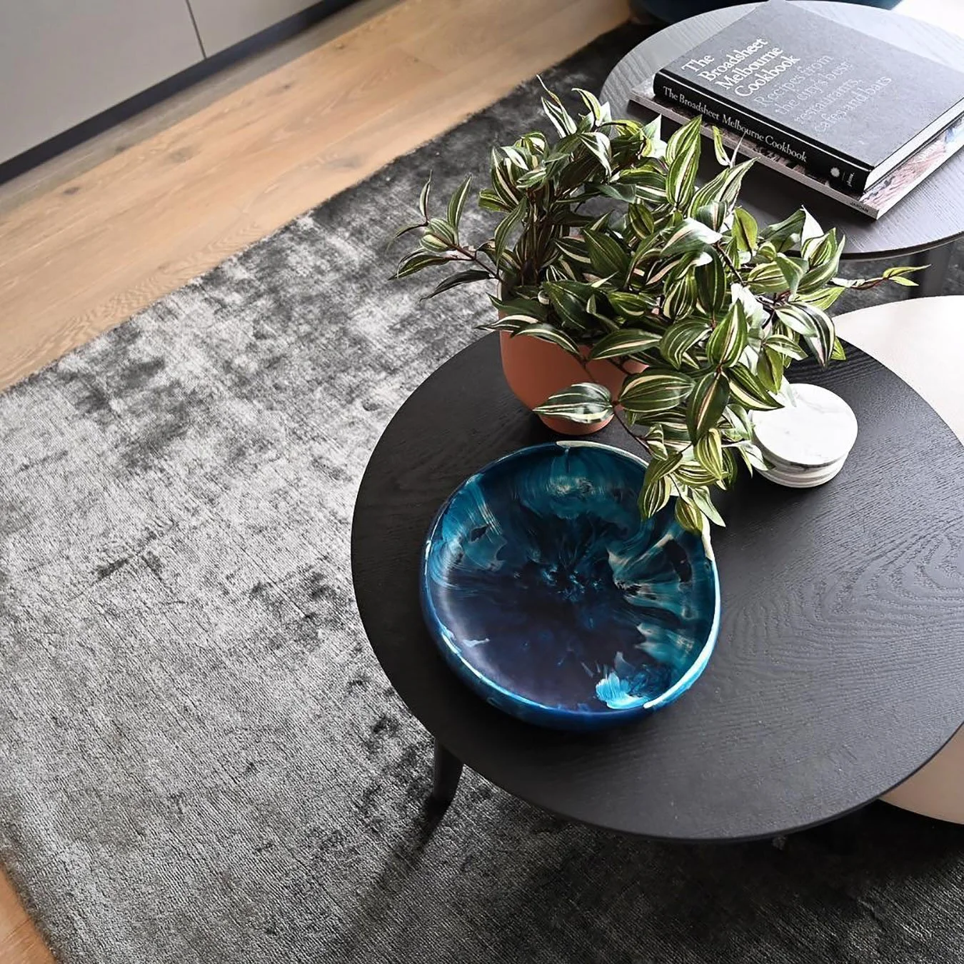A black round coffee table with a potted plant, a blue decorative bowl, a stack of small white plates, and a few books on top. The table is placed on a gray rug with a patterned design, beside a light wooden floor.