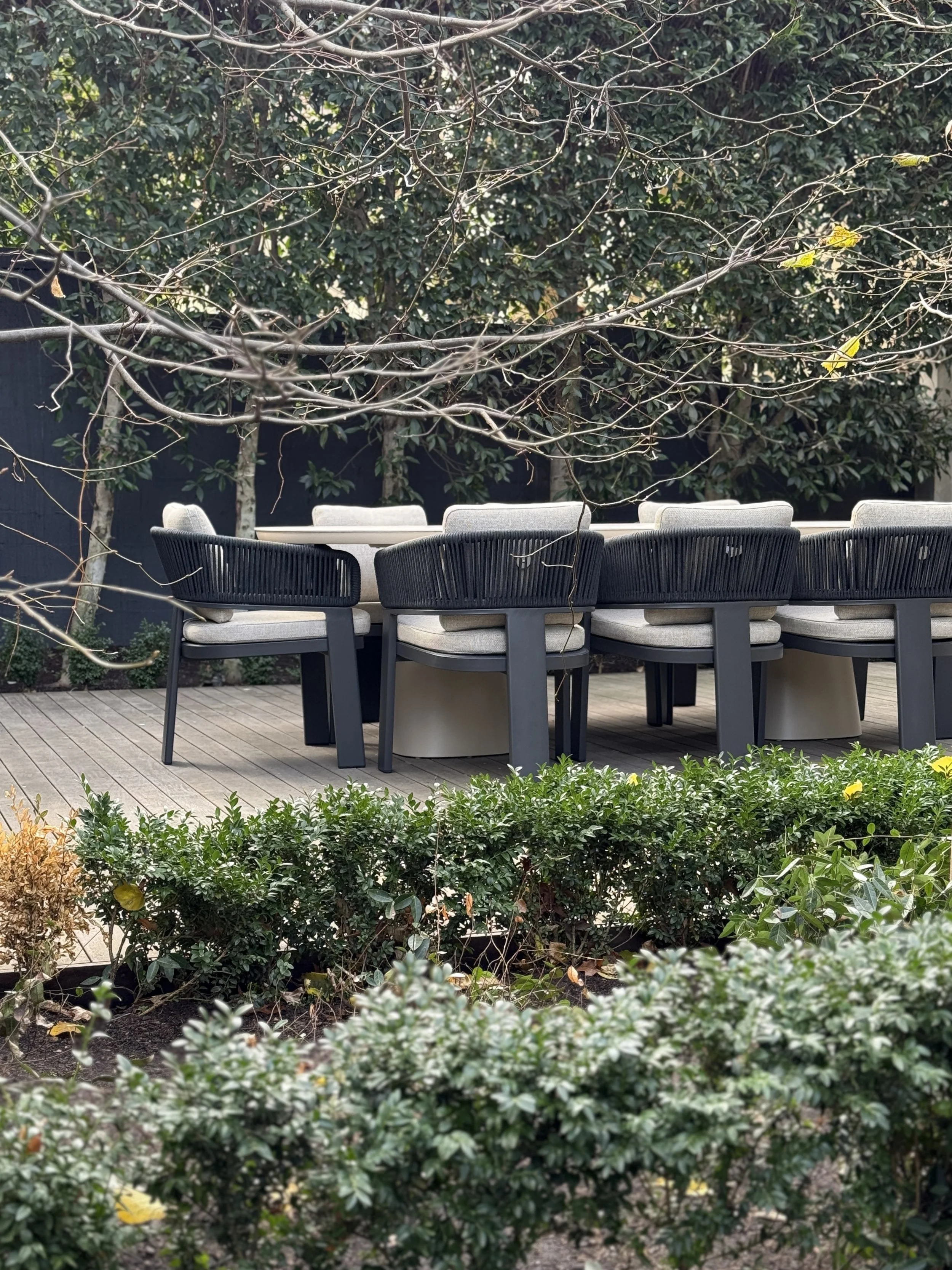 Outdoor patio with a wooden deck featuring a dining table and chairs, surrounded by bushes and trees with some bare branches and a few small green leaves.