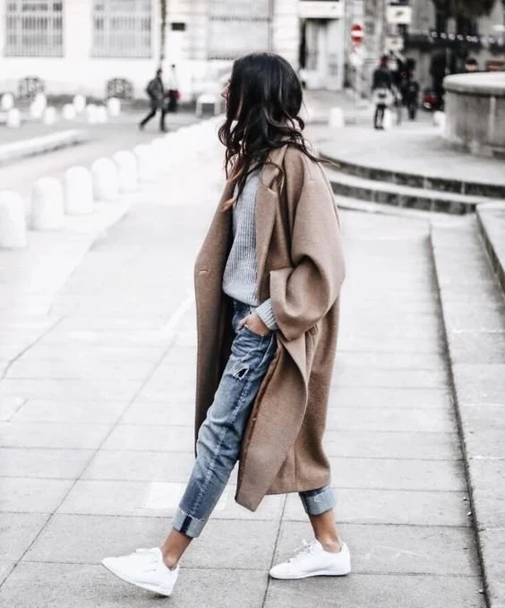 Person wearing a long neutral coat, jeans and white sneakers outdoors.