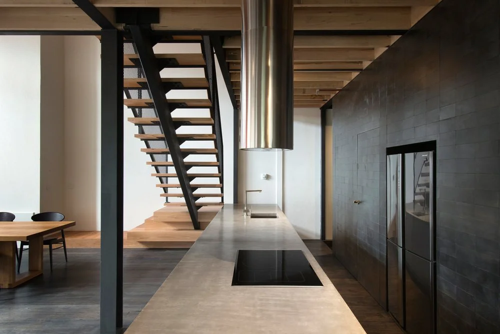 Long kitchen island beneath an open timber staircase and suspended rangehood.