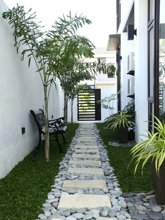Stone path through a narrow garden beside white walls and leafy plants.