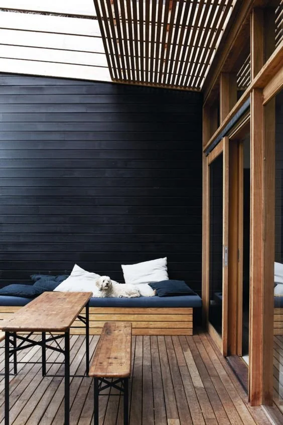 Timber deck and outdoor table beside dark cladding and tall timber doors.
