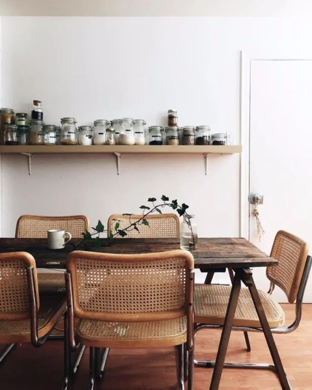 Dining table with timber chairs beneath open shelving of ceramic vessels.