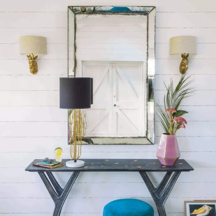 Glass console beneath a tall mirror with dark-shaded lamp and pink vase.