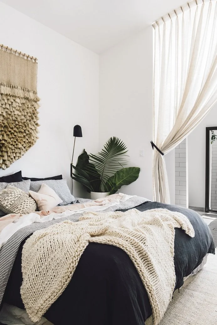 Layered pale linen on a bed beside a woven pendant and softly draped curtains.