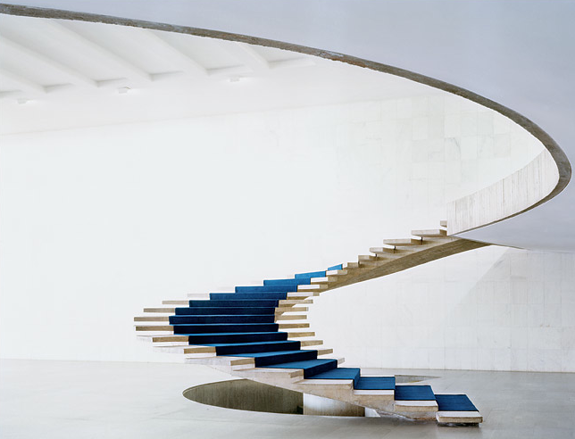 Sweeping white staircase with slender timber steps and blue seating below.