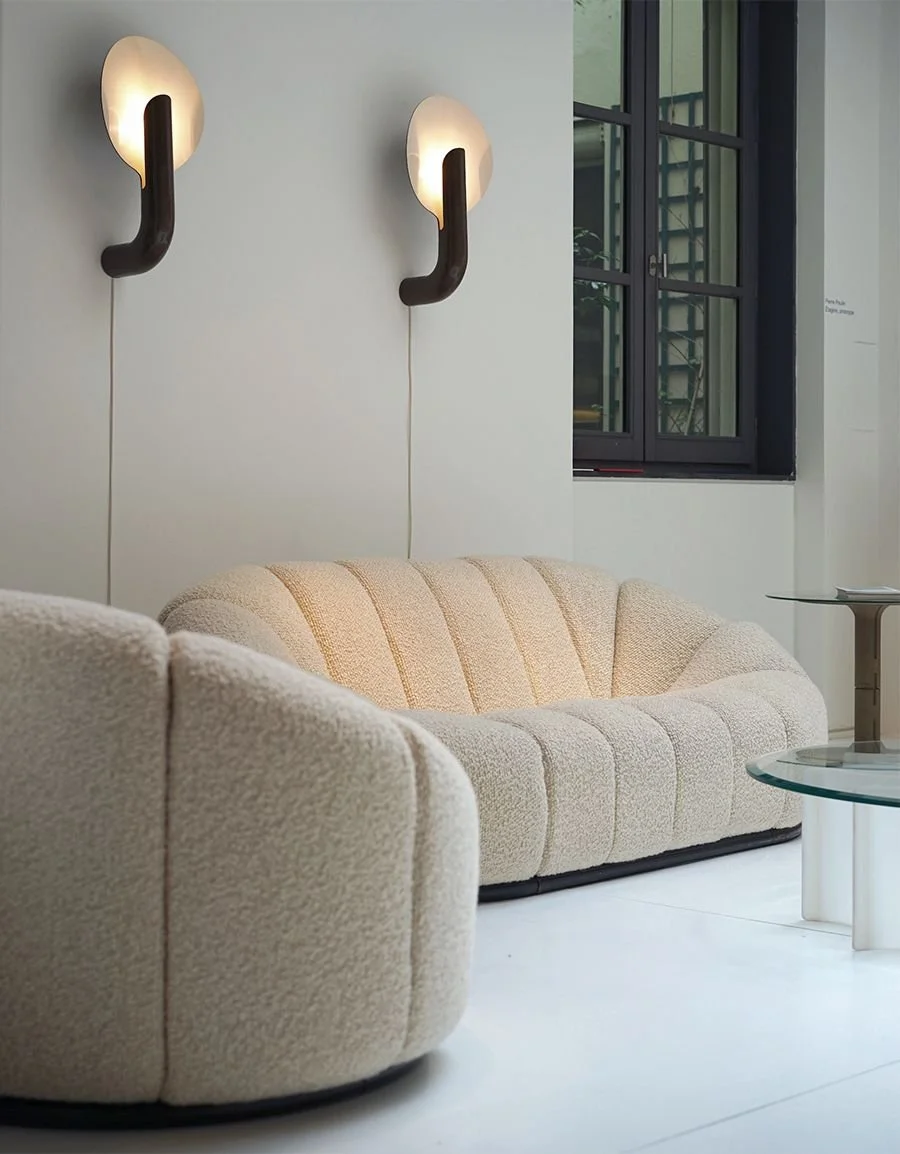 Curved pale sofa beneath twin wall lights beside a dark-framed window.