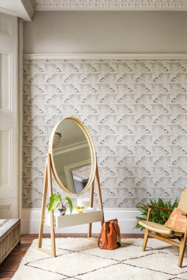 Light patterned wallpaper behind an oval floor mirror and woven chair.
