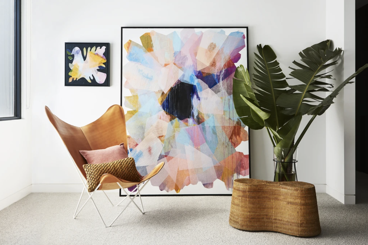 Colourful abstract artwork displayed beside a tan chair and broad-leaf plant.