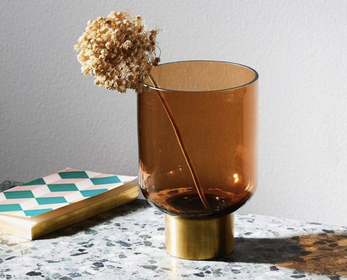 Amber glass vessel with a brass-toned base and dried flower on veined stone.