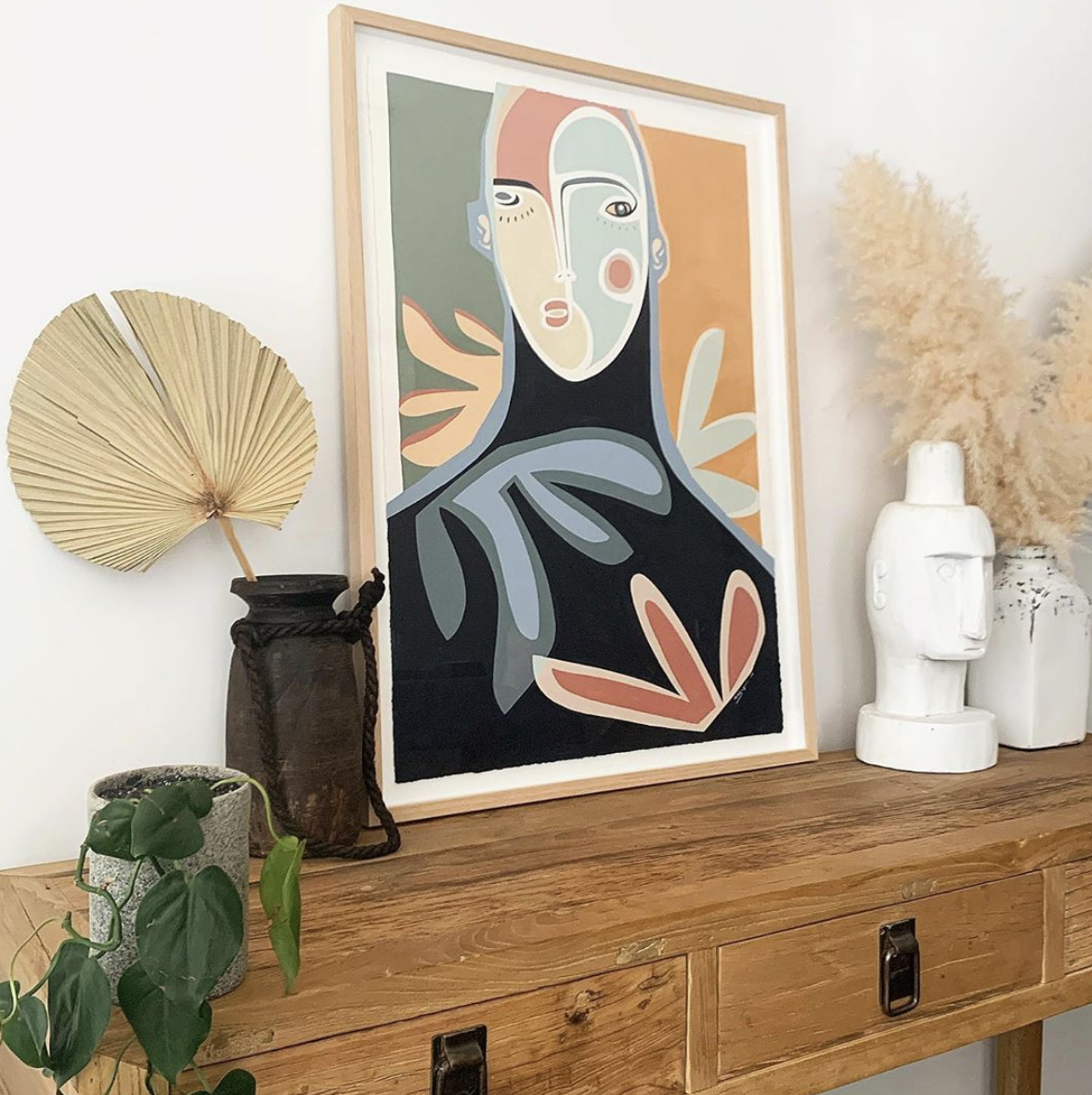 Framed stylised portrait on a timber cabinet beside sculptural vessels and dried foliage.