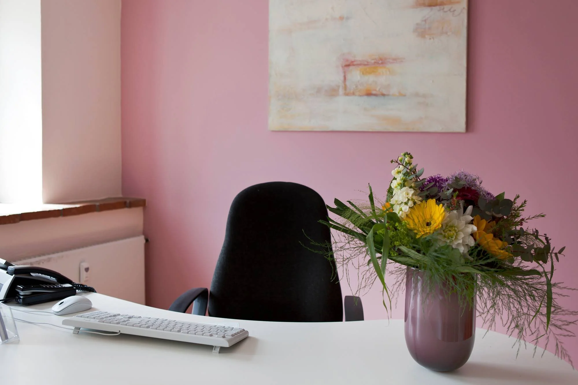 Pink-walled workspace with a dark chair, flowers and a pale desk.