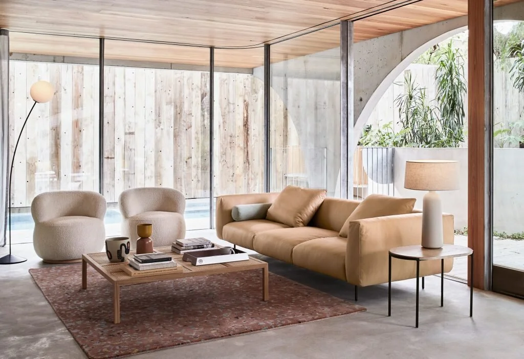 Tan seating and round chairs in a glazed room overlooking trees.