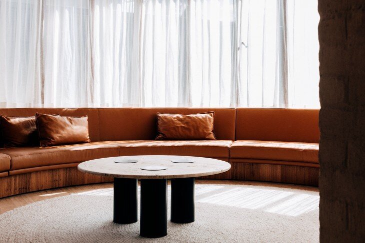 Curved rust-toned sectional sofa and a round coffee table beside sheer curtains.