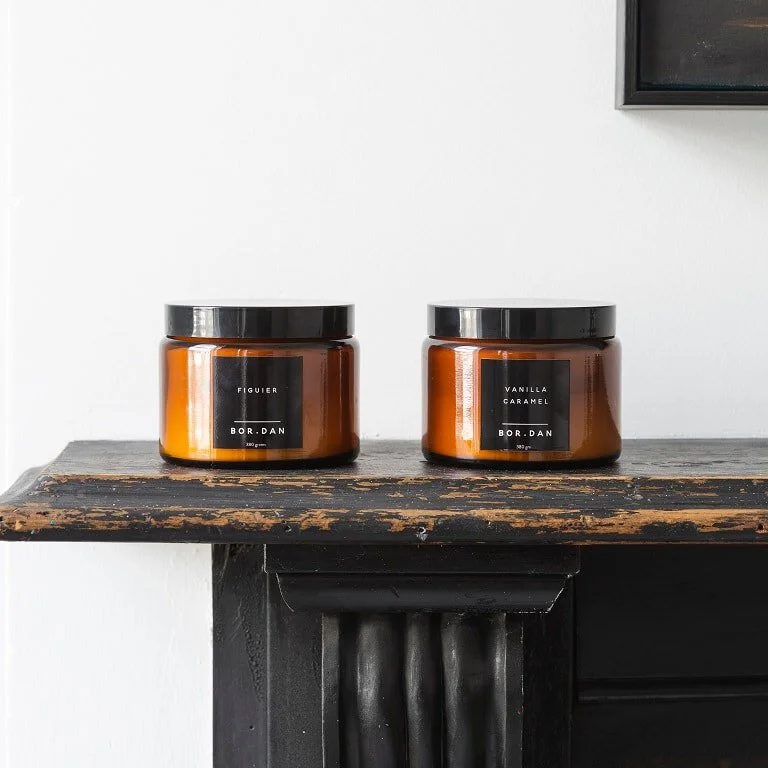 Two amber candle jars with dark lids on a timber mantel.