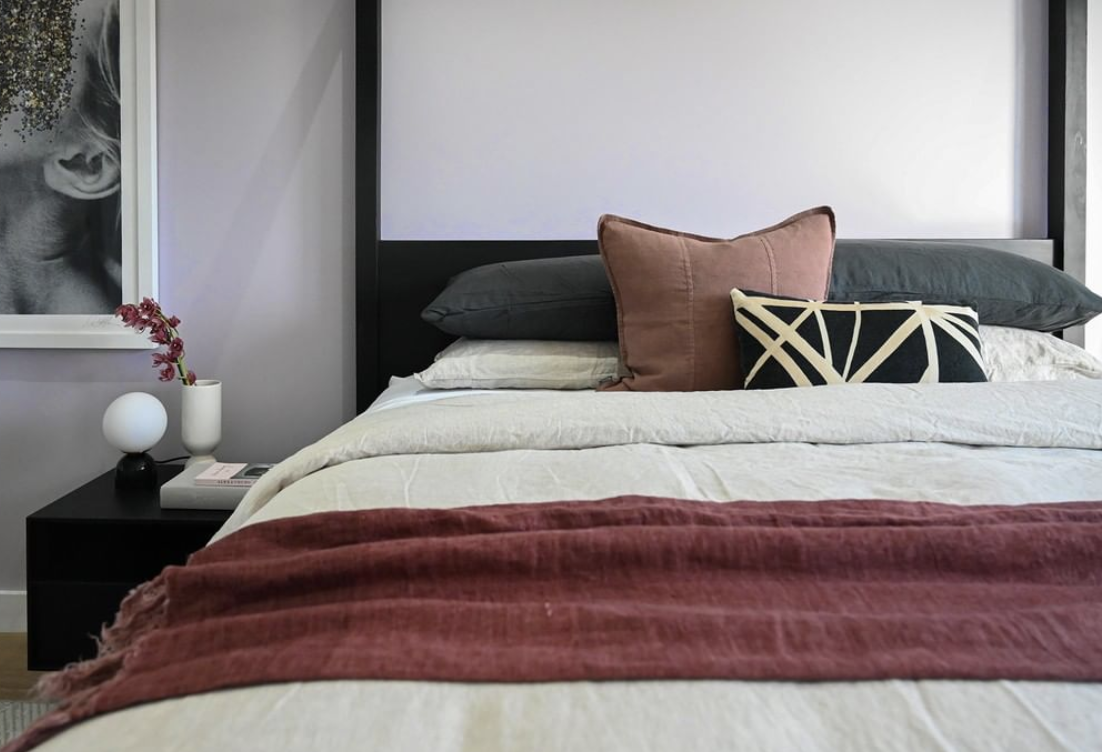 Layered pale bedding with rust-toned textiles and patterned cushions against a dark bedhead.