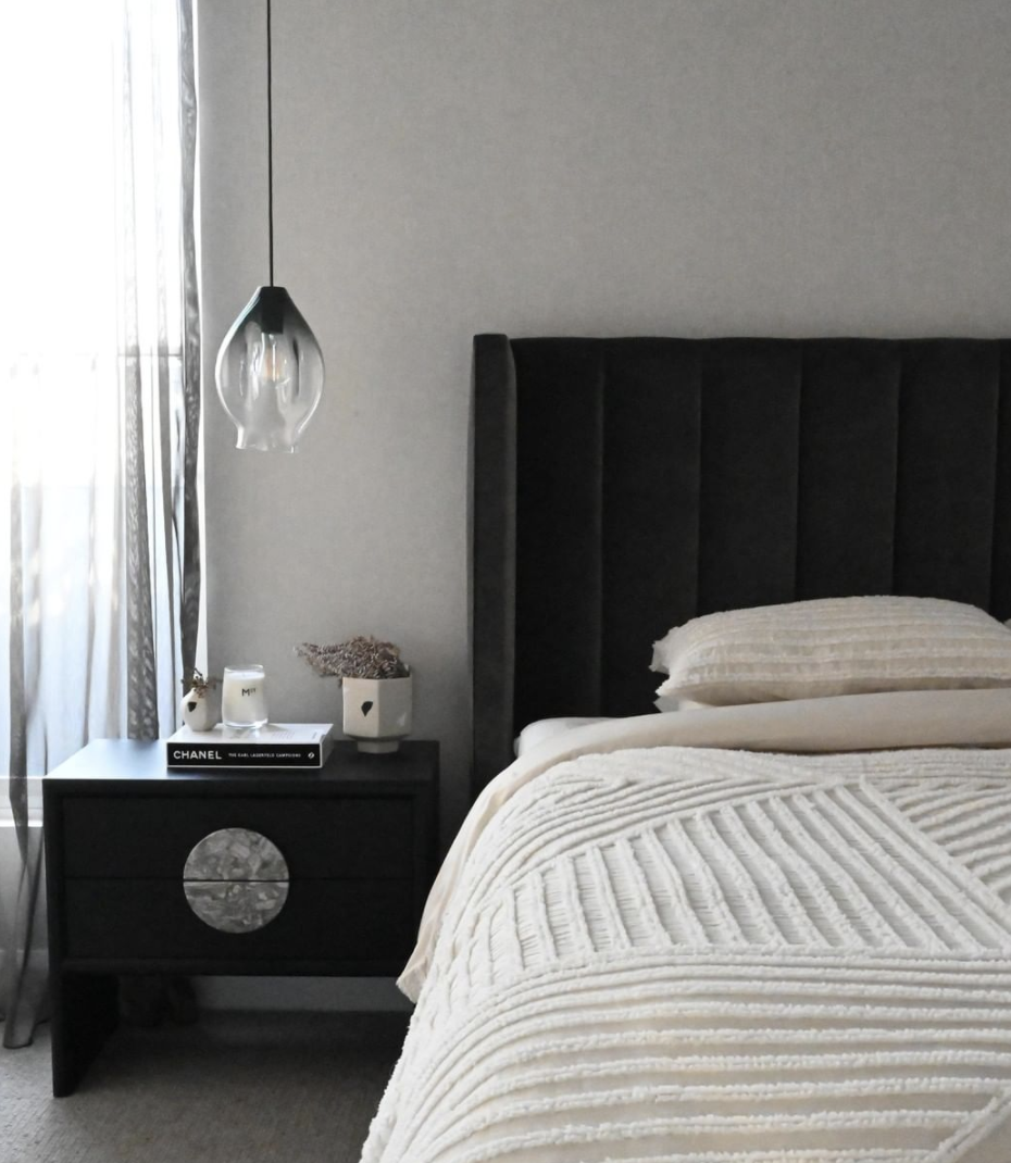 Pale striped bed linen against a dark bedhead beside a glass pendant and black cabinet.
